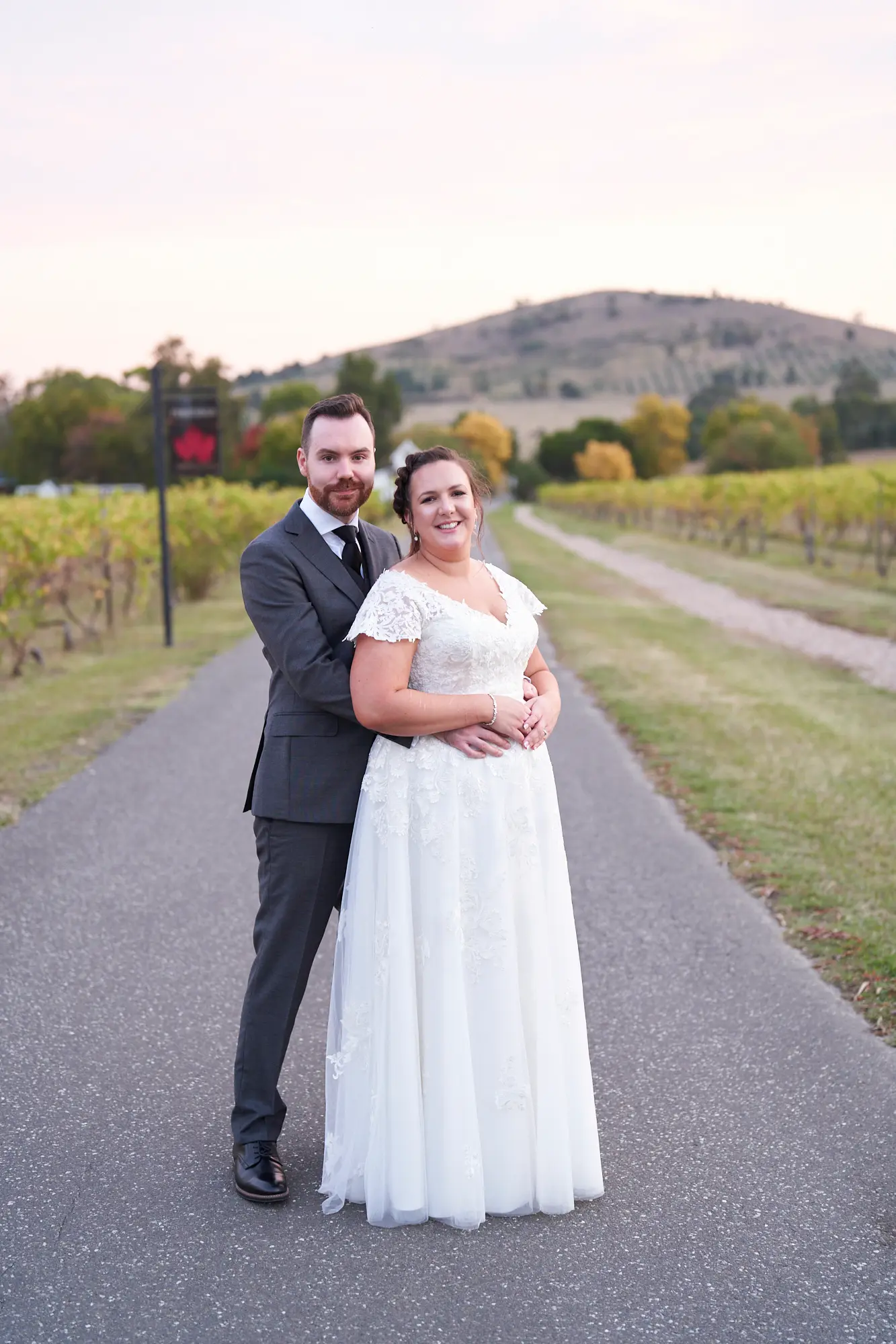 erin jesse joyful embrace driveway fergusson winery yarra valley wedding This wedding photo captures the bride and groom in a joyful, intimate embrace on Fergusson Winery’s scenic driveway in the Yarra Valley. The groom stands behind the bride, arms around her waist, both smiling warmly at the camera. Autumn-toned grapevines frame the paved driveway, with rolling hills and scattered trees in the distance. A rustic wooden winery sign adds charm, while the soft natural light highlights the couple’s happiness. This portrait perfectly blends romance, elegance, and the serene beauty of the vineyard, capturing a heartfelt moment of love and celebration on their special day.
