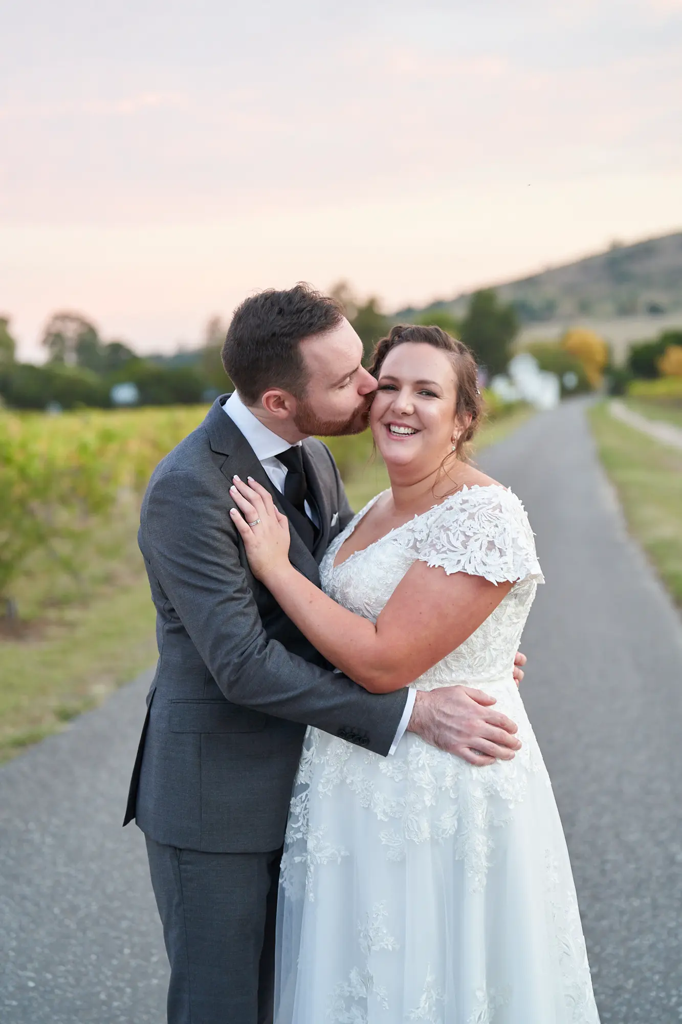 erin jesse kissing driveway vineyard view fergusson winery yarra valley wedding This wedding photo captures the bride and groom sharing a tender kiss on the paved driveway of Fergusson Winery in the Yarra Valley during their portrait session. They stand close, the groom’s arm around the bride’s waist and her hands on his cheeks, fully engrossed in each other. Rows of autumn-toned grapevines line the driveway, with a white guesthouse and rolling hills in the mid- and far-background under a soft evening sky. Surrounded by scattered trees and serene natural beauty, the image conveys intimacy, deep love, rustic elegance, and a tranquil, joyful moment for the newlyweds.