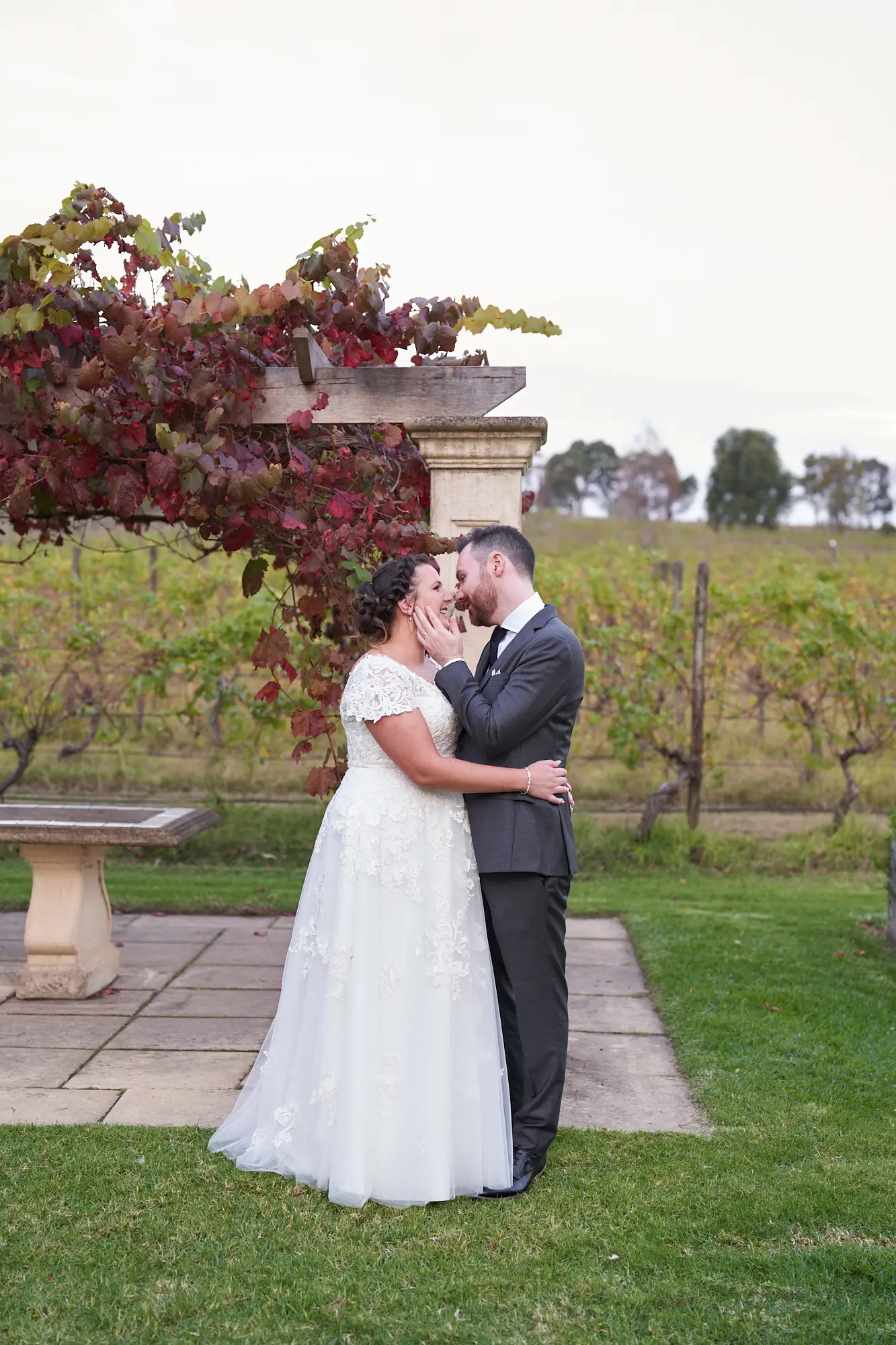 erin jesse romantic close embrace sandstone gazebo fergusson winery yarra valley wedding This wedding photo captures a romantic and intimate moment between the bride and groom at Fergusson Winery in the Yarra Valley. Standing under the "Sandstone Gazebo" draped with red and green autumn grapevines, they are in a close embrace, with the groom’s arm around the bride’s waist and her hands gently on his chest, leaning in toward each other. Rows of vineyards and rolling hills stretch into the background, while manicured lawns and rustic stone elements frame the couple. The portrait conveys emotional connection, warmth, and serene elegance, capturing a private, heartfelt moment before the ceremony.