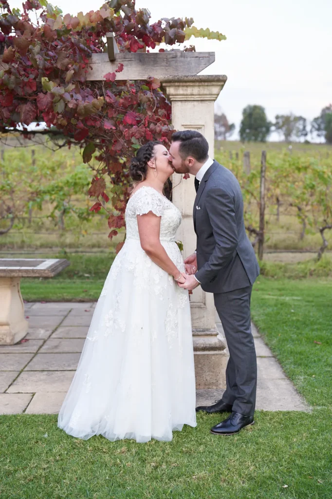 erin jesse romantic gaze under pergola fergusson winery yarra valley wedding This wedding photo captures an intimate, cinematic moment between the bride and groom at Fergusson Winery in the Yarra Valley. Standing under a sandstone pergola draped with vibrant red and green autumn grapevines, they share a gentle kiss, fully absorbed in each other. The groom’s arm wraps around the bride’s waist, her hands resting tenderly on his cheeks. Behind them, rows of lush vineyards stretch into the rolling hills, while the manicured lawn and rustic stone elements frame the scene. The portrait exudes romantic elegance, deep emotional connection, and serene, timeless beauty in a picturesque vineyard setting.