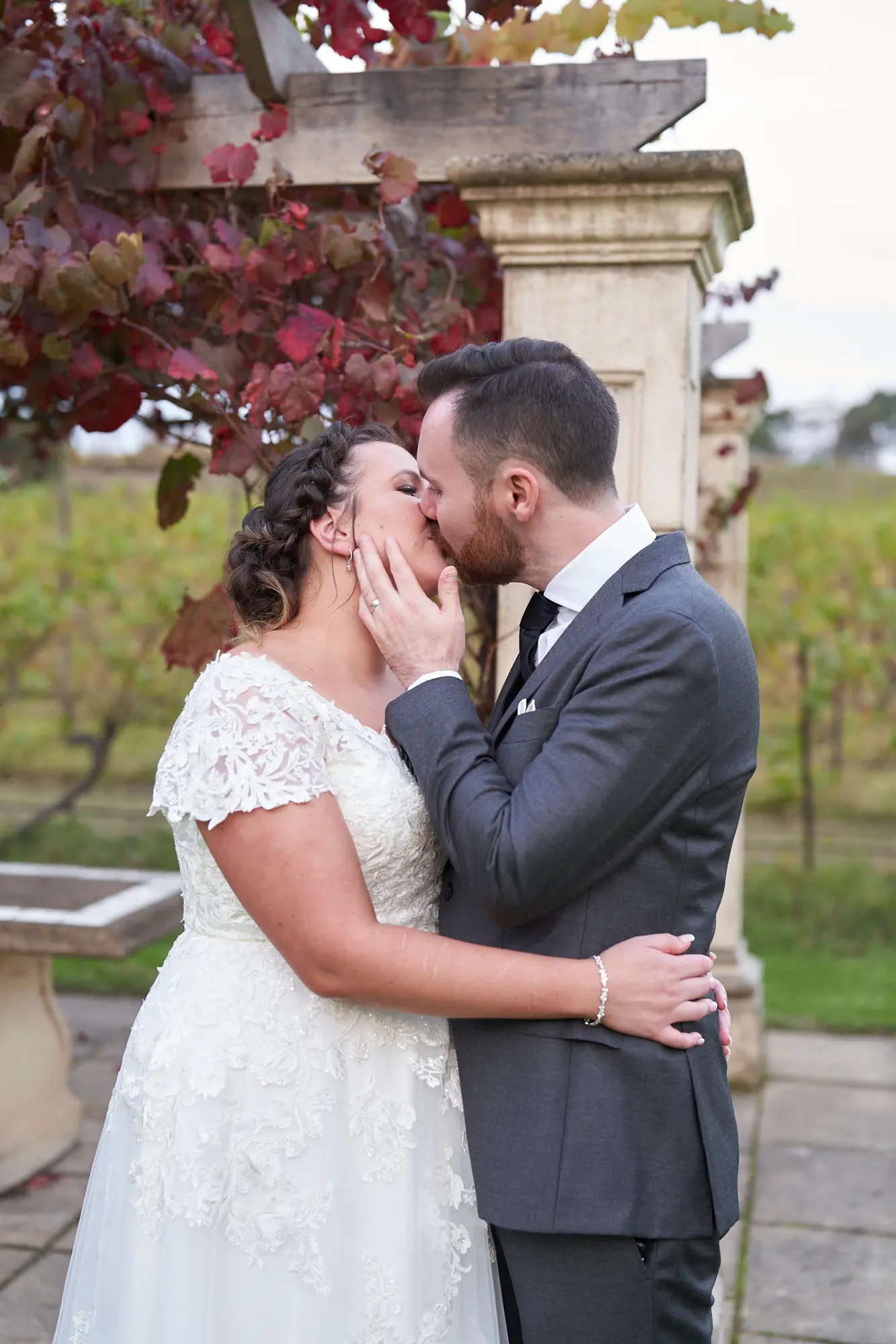 erin jesse romantic kiss sandstone gazebo fergusson winery yarra valley wedding This wedding photo captures an intimate and romantic moment as the bride and groom share a gentle kiss under a vine-covered sandstone gazebo at Fergusson Winery in the Yarra Valley. Embraced closely, with the groom’s arm around the bride’s waist and her hands on his cheeks, they are framed by autumn-toned grapevines. The lush vineyards and rolling hills in the background add depth, creating a serene, heartfelt, and joyful portrait that blends rustic elegance with natural beauty, highlighting their love and private connection during their outdoor wedding portrait session.