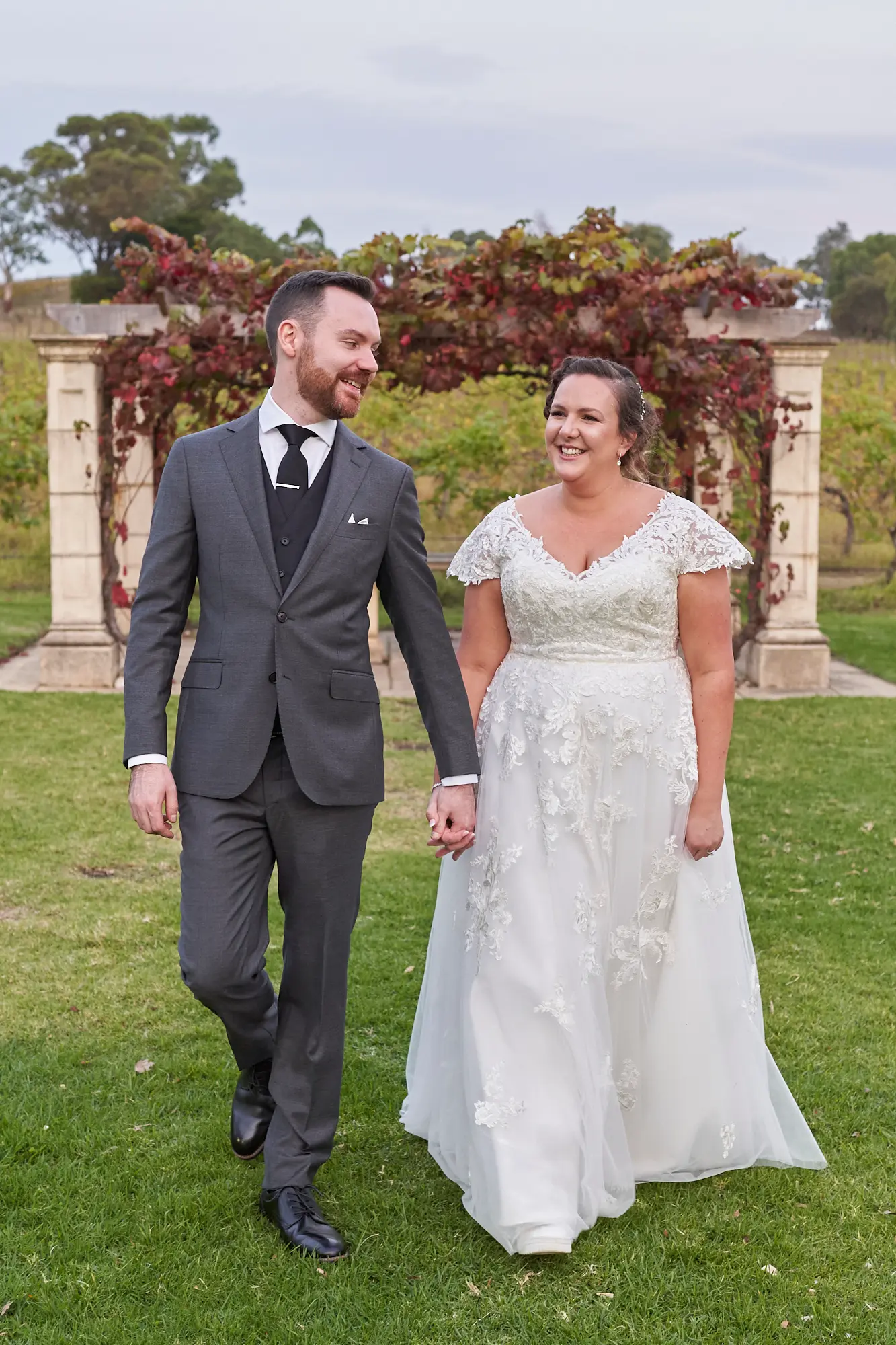 erin jesse walking hand in hand sandstone gazebo fergusson winery yarra valley wedding This wedding photo captures a tender, joyful moment as the bride and groom walk hand-in-hand across the green lawn at Fergusson Winery in the Yarra Valley. They smile warmly at each other, fully immersed in their shared happiness and connection. Framed by a sandstone gazebo draped in vibrant autumn grapevines and surrounded by rows of lush vineyards, the scene combines natural beauty and rustic elegance. Their relaxed, confident stroll reflects the love and excitement of their wedding day, creating a serene, storytelling portrait that showcases both their intimacy and the picturesque winery setting.