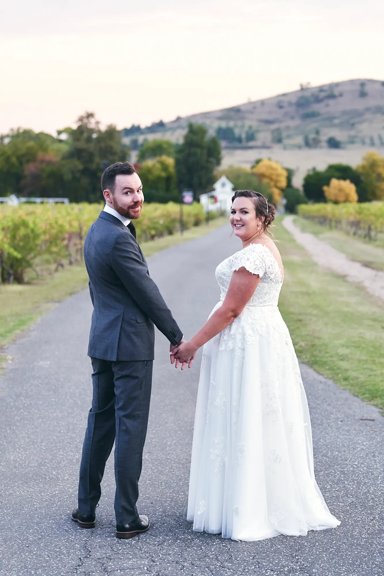 erin jesse walking hand in hand vineyard driveway fergusson winery yarra valley wedding This wedding photo captures the bride and groom walking hand-in-hand down a paved vineyard driveway at Fergusson Winery in the Yarra Valley, looking back over their shoulders with joyful, wide smiles. The groom wears a dark gray suit and the bride a white lace dress, as autumn-toned grapevines line both sides. In the mid-background, a charming white guesthouse with a red roof peeks through, and rolling hills frame the scene under a bright sky. The image exudes warmth, rustic elegance, and a serene, romantic moment, highlighting the couple’s happiness and shared excitement for their future together.