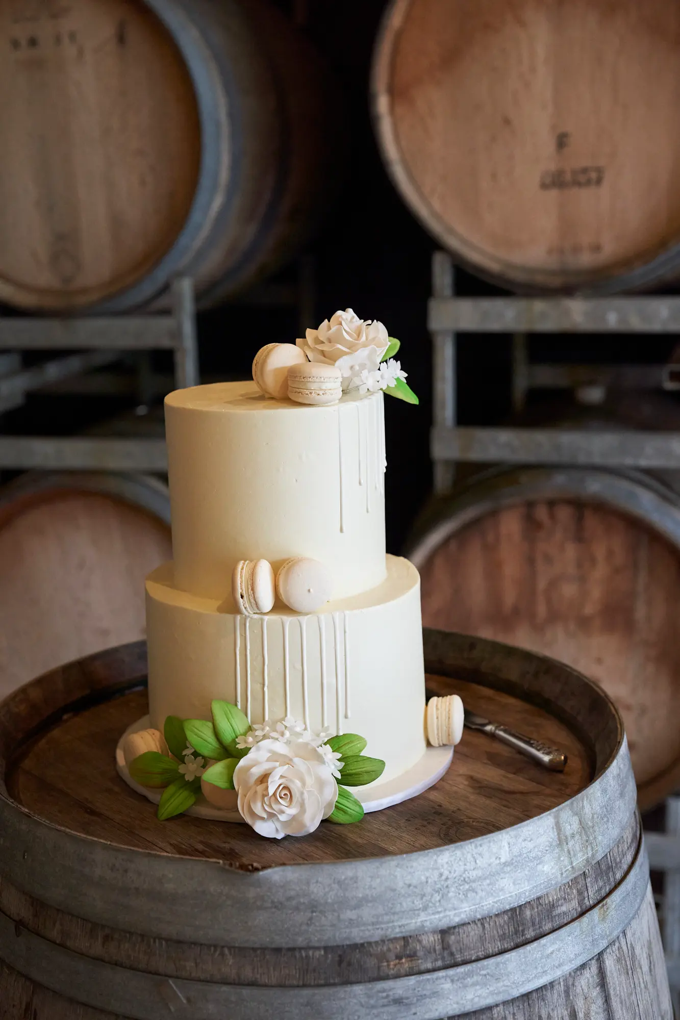 erin jesse wedding cake barrel room fergusson winery This photo showcases a two-tiered wedding cake elegantly displayed on a rustic wooden wine barrel in the barrel room at Fergusson Winery. The ivory buttercream cake features a gentle drip effect and is adorned with white macarons and realistic sugar roses with green leaves. Behind, stacked wooden barrels add warm, textured depth, highlighting the cozy, rustic elegance of the setting. Soft lighting illuminates the cake, emphasizing its delicate details, while the prepared barrel and knife suggest the impending joyful moment when the bride and groom will share their first ceremonial cut during their intimate, elegant reception.