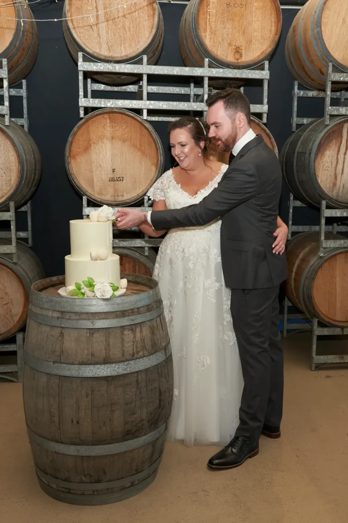 erin jesse wedding cake cutting fergusson winery This wedding photo captures a joyful and intimate moment as the bride and groom make the first cut of their two-tiered wedding cake in the barrel room at Fergusson Winery. Standing closely together, the groom’s arm around the bride’s waist, both smile with delight at this shared milestone. The warm, rustic backdrop of stacked wooden wine barrels, with authentic stenciled markings, adds texture and character to the scene. The cozy atmosphere highlights the couple’s connection and the celebratory joy of the reception, blending rustic charm with elegance in a heartfelt, memorable moment.