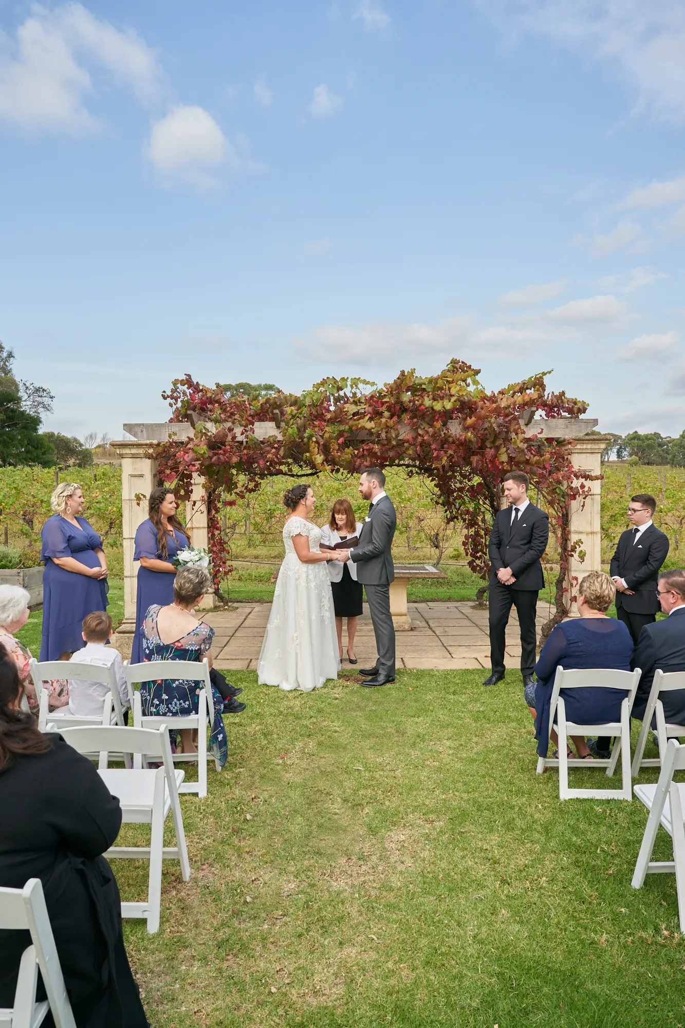 erin jesse wedding ceremony vows fergusson winery yarra valley Outdoor wedding ceremony at Fergusson Winery in the Yarra Valley, showing the bride and groom standing beneath a vine-covered pergola as they exchange vows before their officiant. Bridesmaids in blue dresses and groomsmen in suits stand on either side, while guests seated on white chairs watch from the lawn. The rustic pergola draped in red and green autumn grapevines frames the couple, with rows of vineyards and a bright sky behind them, creating a serene, elegant, and joyfully intimate moment during the exchange of vows.