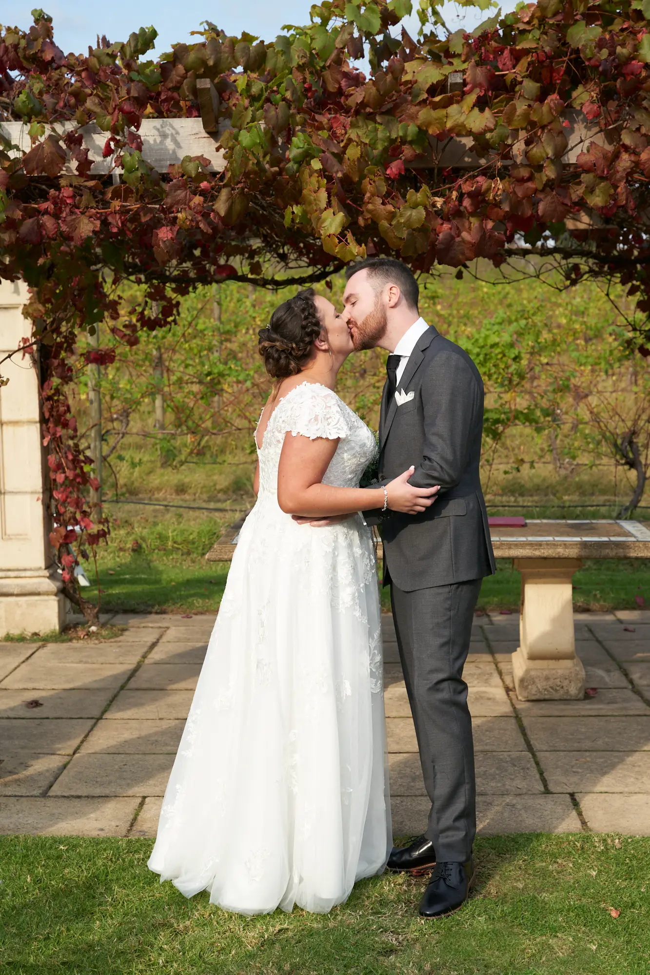 erin jesse wedding kiss sandstone gazebo fergusson winery yarra valley Bride and groom sharing a gentle, romantic kiss beneath the vine-covered Sandstone Gazebo at Fergusson Winery in the Yarra Valley. Embracing closely, the groom holds the bride around her waist while she cups his cheeks, creating an intimate and emotional moment. Lush grapevines with vibrant red and green autumn leaves frame the couple, with rows of vineyards stretching into the distance under bright natural light. This serene, heartfelt scene captures the joy, rustic elegance, and idyllic beauty of their outdoor wedding celebration.