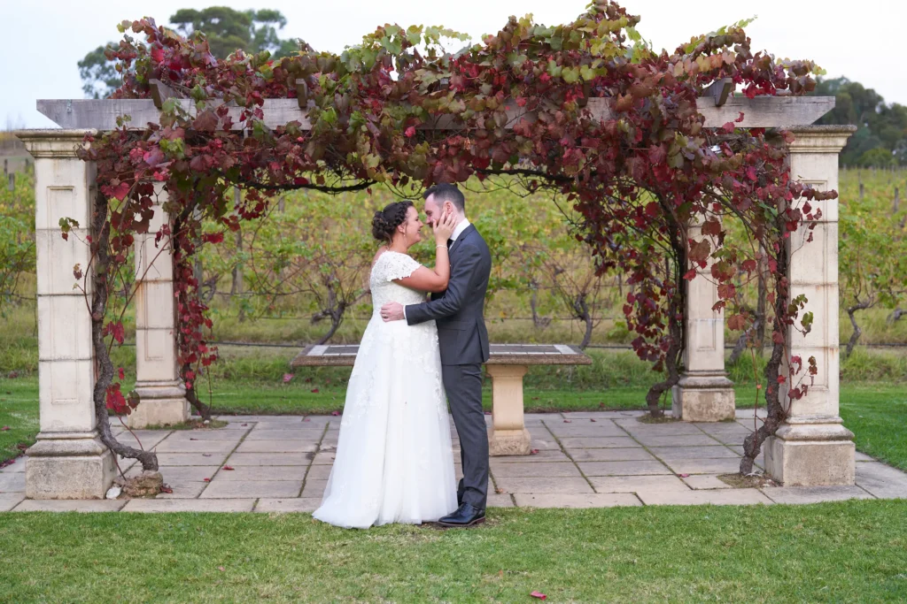 erin jesse wedding portrait fergusson winery yarra valley pergola Erin and Jesse share a tender, intimate moment under a vine-covered pergola at Fergusson Winery in the Yarra Valley, with the groom holding the bride close around her waist and her hands resting gently on his cheeks. Surrounded by autumn-red and green grapevines, manicured lawns, and rows of rolling vineyards stretching into the horizon, this romantic wedding portrait captures their deep love, joyful connection, and the serene, rustic elegance of the winery, perfectly blending the couple’s heartfelt emotion with the natural beauty and charm of this iconic vineyard setting.