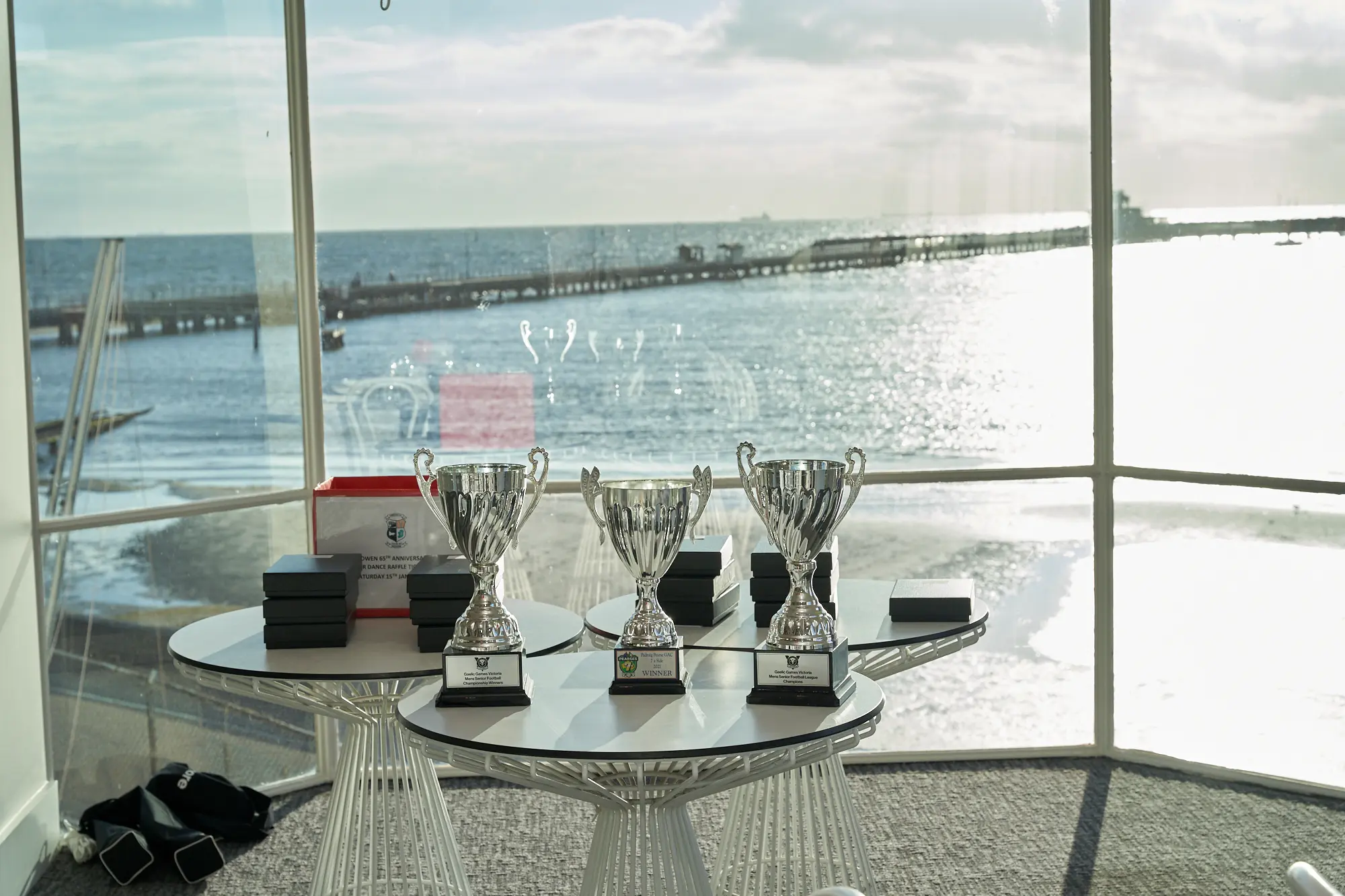 garryowens 65th anniversary ball awards trophies display waterfront A beautifully arranged awards display awaits the Garryowen’s 65th Anniversary Ball, featuring three polished silver trophies placed on two modern white round tables with ribbed transparent bases. Behind them, neatly stacked black and red presentation boxes hint at the upcoming ceremony. Large windows frame a stunning coastal view with sparkling water and a long pier stretching into the bay. Bright natural light fills the spacious venue, highlighting the elegant décor and creating an atmosphere of anticipation and prestige for the evening’s awards night celebration.