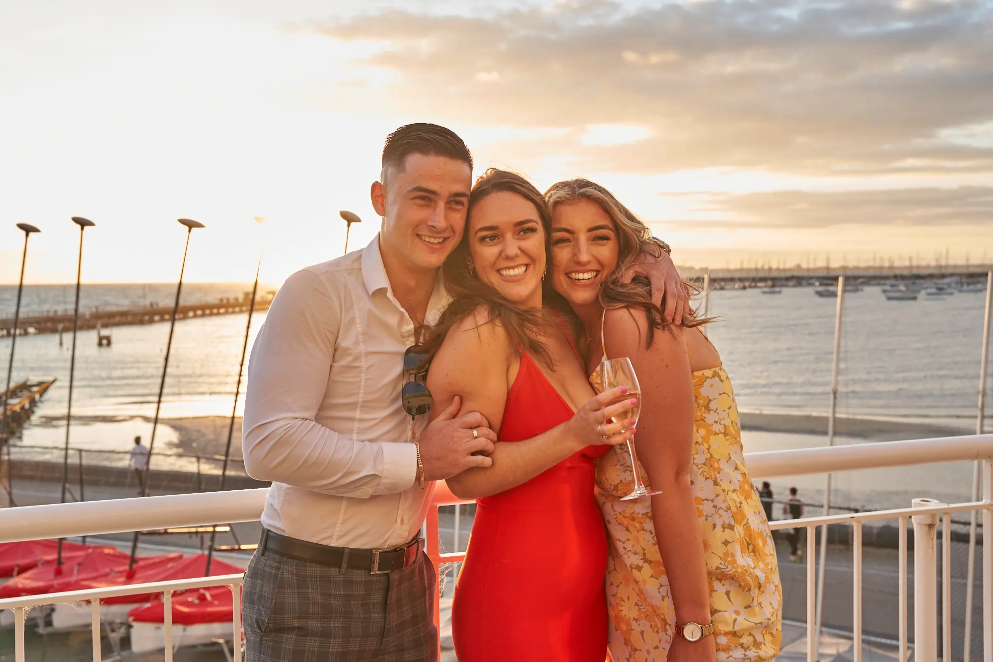 garryowens 65th anniversary ball candid balcony portrait Three guests pose candidly on a balcony during Garryowen’s 65th Anniversary Ball at the Harbour Room, Royal Melbourne Yacht Squadron. A man in formal attire has his arm around a woman in a red dress, who links arms with another woman in a yellow floral dress holding a wine glass. They smile warmly at the camera, set against a golden-hour waterfront view with calm bay waters, distant boats, a long pier, and white balcony railings. The scene radiates festive, relaxed elegance, capturing a joyful, intimate moment of camaraderie and natural celebration at the prestigious gala.