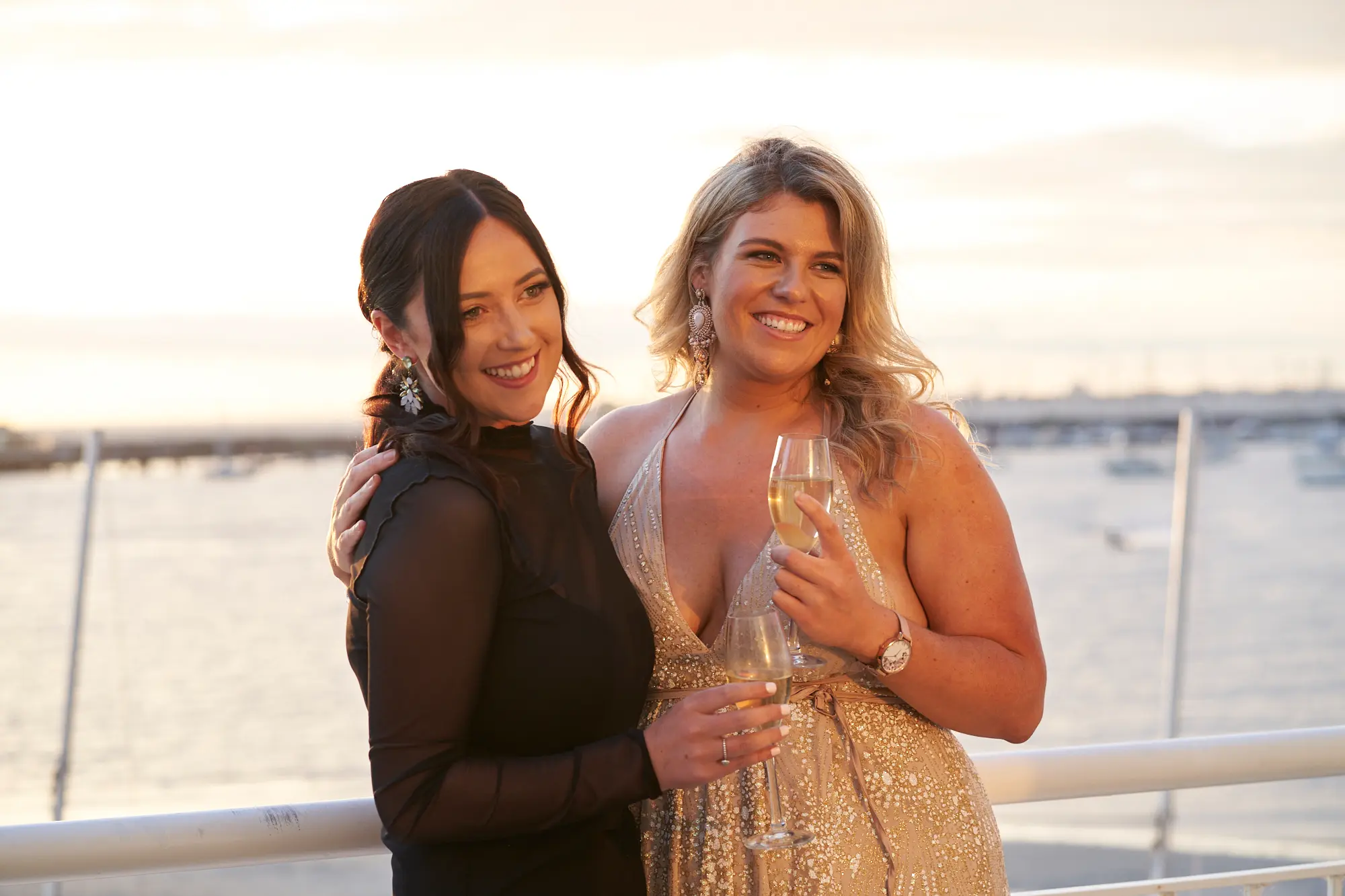 garryowens 65th anniversary ball candid golden hour duo Two elegantly dressed women pose closely on an outdoor deck during Garryowen’s 65th Anniversary Ball at the Harbour Room, Royal Melbourne Yacht Squadron. One wears a black dress with her arm around the other in a sparkling gold gown holding a champagne glass. They smile warmly, framed by white balcony railings and a golden-hour waterfront view of St Kilda Pier and Port Phillip Bay, creating a joyful, sophisticated, and celebratory atmosphere.