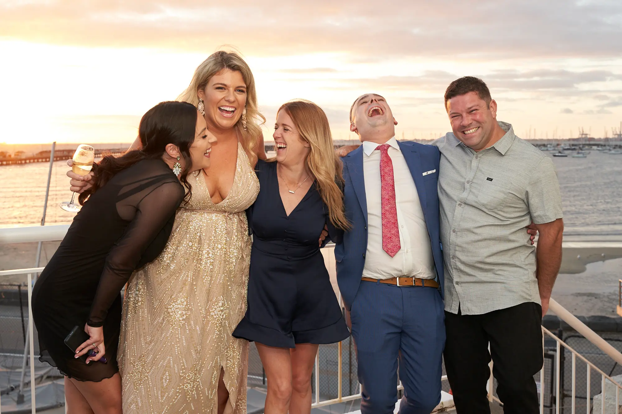 garryowens 65th anniversary ball candid golden hour group five A group of five elegantly dressed guests—three women and two men—share a candid, joyful moment on an outdoor balcony during Garryowen’s 65th Anniversary Ball at the Harbour Room, Royal Melbourne Yacht Squadron. They are laughing, embracing, and raising champagne glasses, radiating warmth and camaraderie. The white balcony railings frame a breathtaking golden-hour view of St Kilda Pier and Port Phillip Bay, with docked boats in the distance. The soft, natural light highlights their formal attire and happy expressions, perfectly capturing the lively, festive, and intimate atmosphere of this celebratory gala evening.