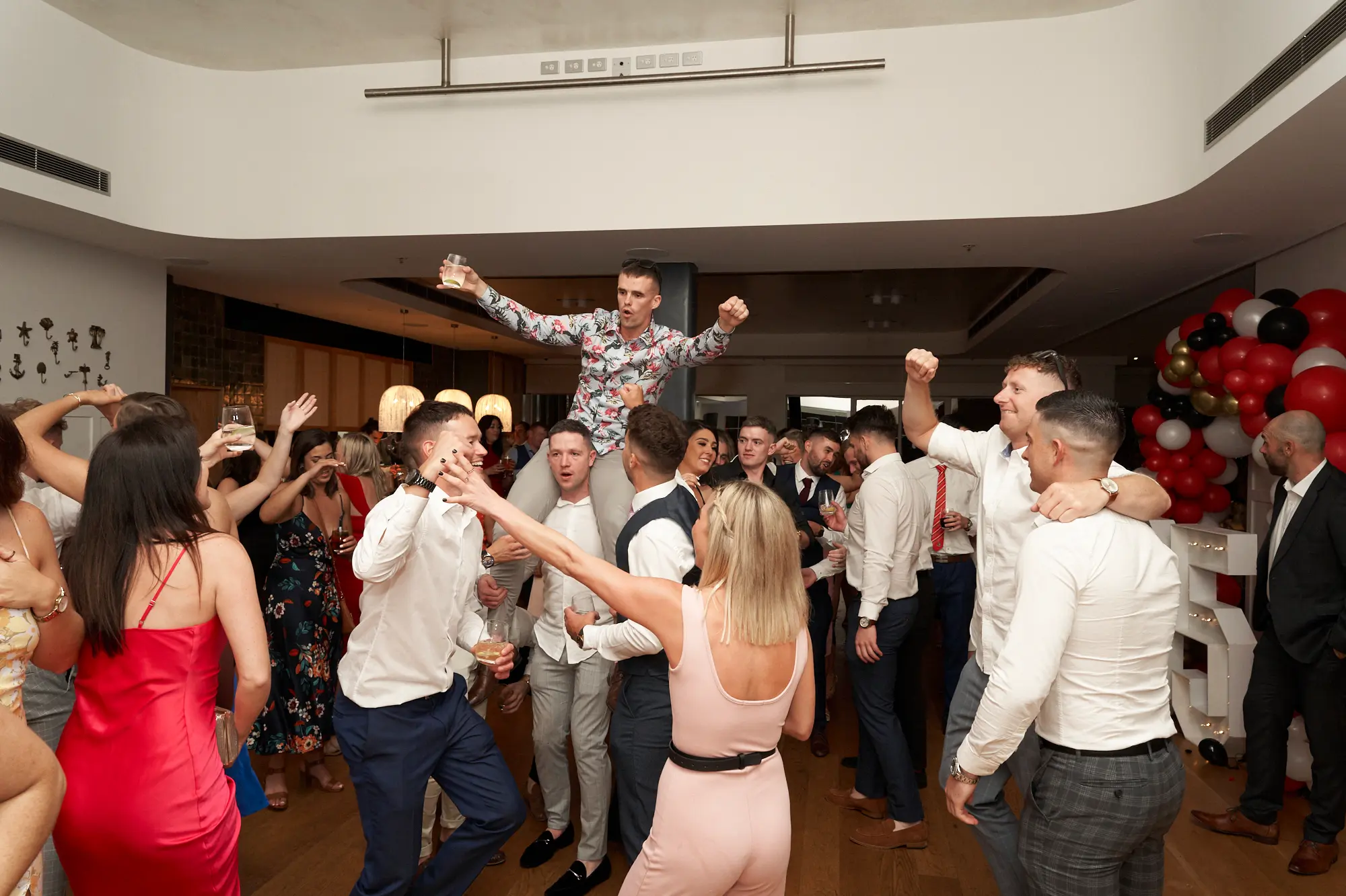 garryowens 65th anniversary ball chairing celebration Guests celebrate energetically at Garryowen’s 65th Anniversary Ball at the Royal Melbourne Yacht Squadron. A man in a patterned shirt is lifted onto another’s shoulders, holding a glass and cheering, while surrounding guests in formal and cocktail attire clap, raise drinks, and laugh. A woman in a red dress is captured mid-cheer. The polished hardwood floor, white walls, festive balloon arch in red, black, gold, and white, and well-lit dining tables create a vibrant, lively party atmosphere, capturing the excitement and camaraderie of a jubilant awards night celebration.