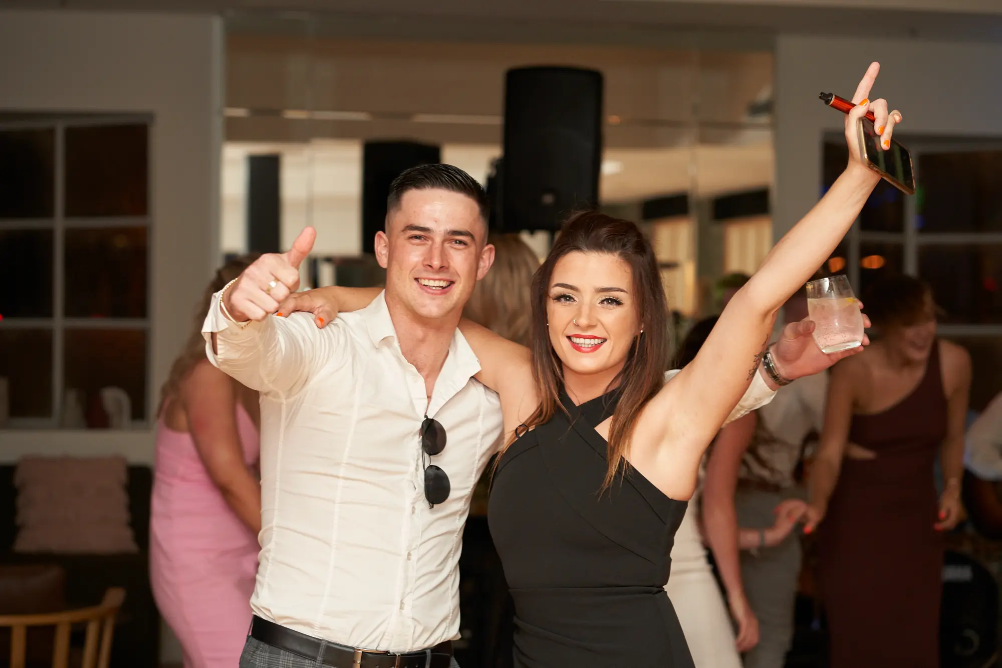 garryowens 65th anniversary ball couple celebration indoor A joyful couple poses at Garryowen’s 65th Anniversary Ball, standing closely together and smiling broadly at the camera. The man has his arm around the woman’s shoulder and gives a thumbs-up while holding a drink. The woman raises her arm excitedly, holding a glass and a small red object. Guests mingle in the softly lit, elegant indoor venue with polished hardwood floors and architectural details, creating a sophisticated ambiance. Their energetic, friendly poses capture a moment of lively celebration, camaraderie, and excitement at a well-attended formal gala.