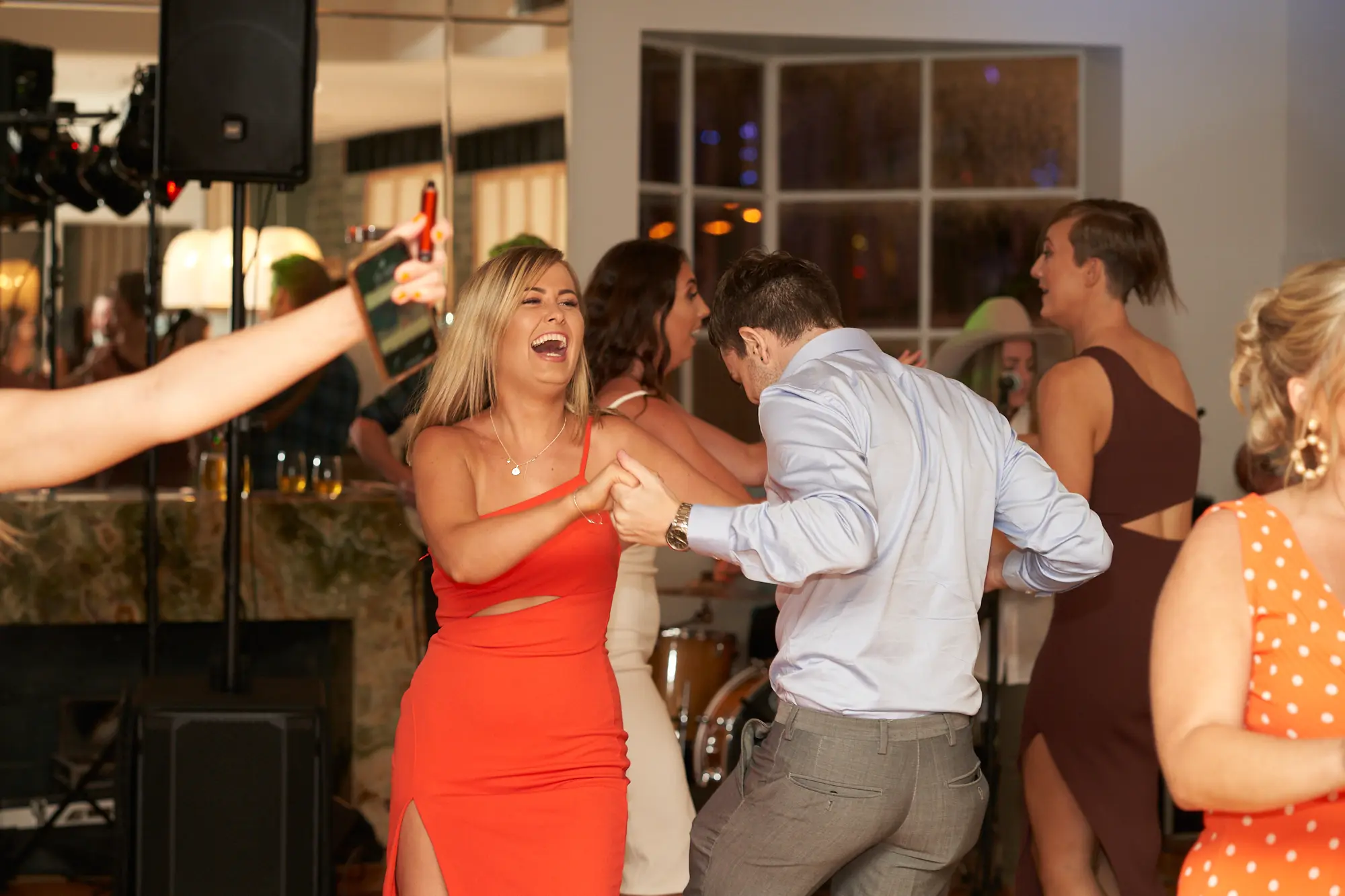garryowens 65th anniversary ball couple dancing laugh A lively dance moment at Garryowen’s 65th Anniversary Ball at the Royal Melbourne Yacht Squadron. A woman in an orange dress laughs joyfully while dancing with a man in a light blue shirt. Nearby, a woman in a maroon dress dances, and a guest on the far left holds up a smartphone, capturing the energetic scene. Other guests mingle and dance across the polished hardwood floor, with mirrored walls multiplying the festive activity and windows casting soft ambient light. A stone fireplace, stage equipment, and a bar area complete the vibrant, high-energy atmosphere of celebration and shared