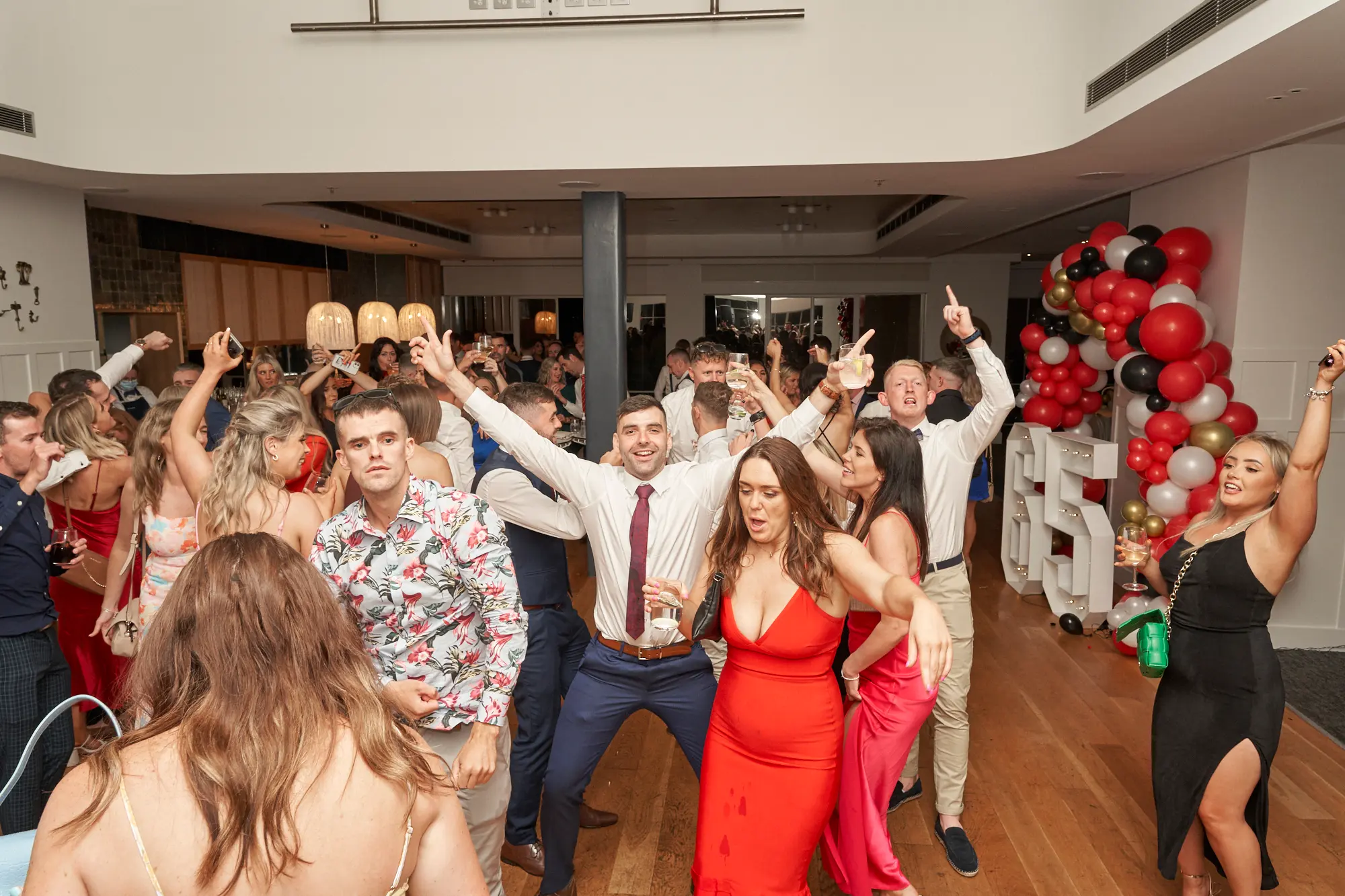 garryowens 65th anniversary ball dance floor celebration wide shot Guests celebrate energetically at Garryowen’s 65th Anniversary Ball at the Royal Melbourne Yacht Squadron. Two men in the center are captured mid-cheer with wide smiles and arms raised, while a woman in red in the foreground and a woman in black to the right also cheer enthusiastically. Guests in formal and cocktail attire hold drinks and interact, adding to the lively, high-energy atmosphere. The polished hardwood floor, white walls, large festive balloon arch in red, black, gold, and white with illuminated numbers, and softly lit dining tables enhance the festive and vibrant party scene of a memorable awards night.