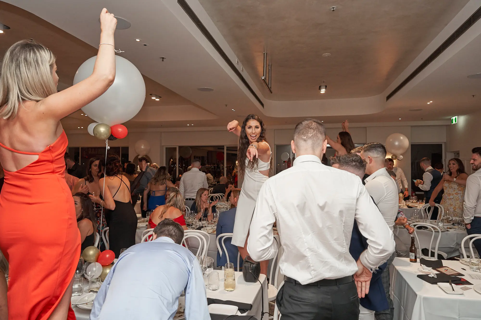 garryowens 65th anniversary ball dance floor celebration Guests revel on the dance floor at Garryowen’s 65th Anniversary Ball at royal Melbourne yacht squadron. A woman in an orange dress dances with arms raised, while another in white stands on a chair, smiling and pointing at the camera. Men in light blue and white shirts engage energetically nearby. The room is filled with mingling guests, festive red, white, gold, and black balloon decorations, and polished hardwood floors. Tables with white linens and chairs frame the scene. The atmosphere is vibrant, joyful, and electric, capturing the high energy, excitement, and shared happiness of a memorable gala celebration.