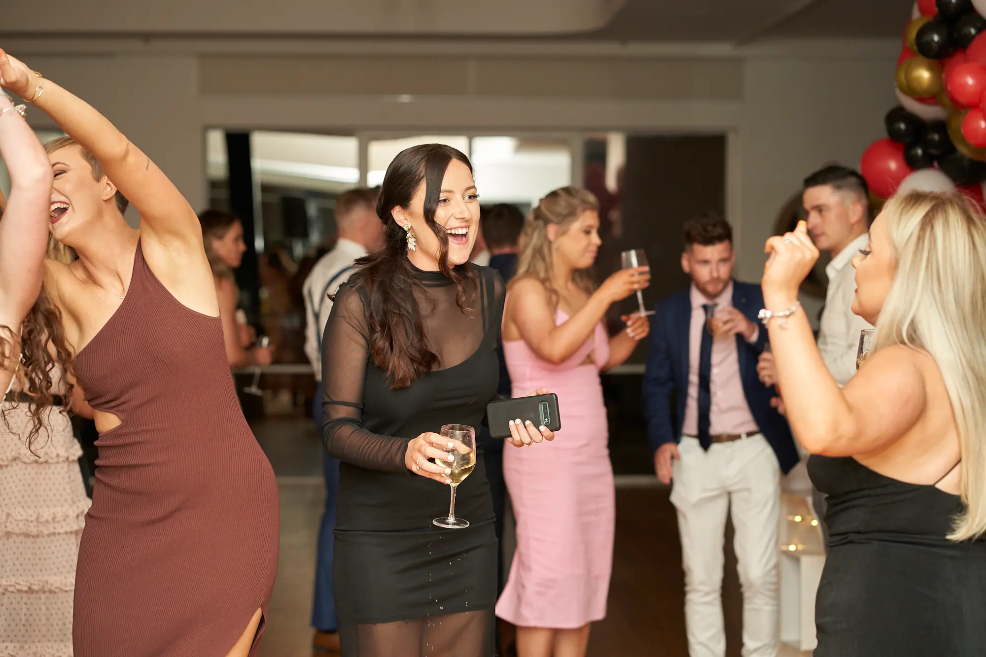 garryowens 65th anniversary ball dancing celebration Friends dancing and celebrating at Garryowen’s 65th Anniversary Ball at the Royal Melbourne Yacht Squadron. Two women in black dresses are laughing and dancing energetically, one holding a glass of wine, while another woman in brown and one in pink join in the lively fun. Other guests mingle in the background near a bar and tables. A large red, black, gold, and white balloon arch adds a festive touch, and polished hardwood floors reflect the warm lighting. The scene captures joyful movement, high energy, and the vibrant, celebratory atmosphere of a memorable awards night.
