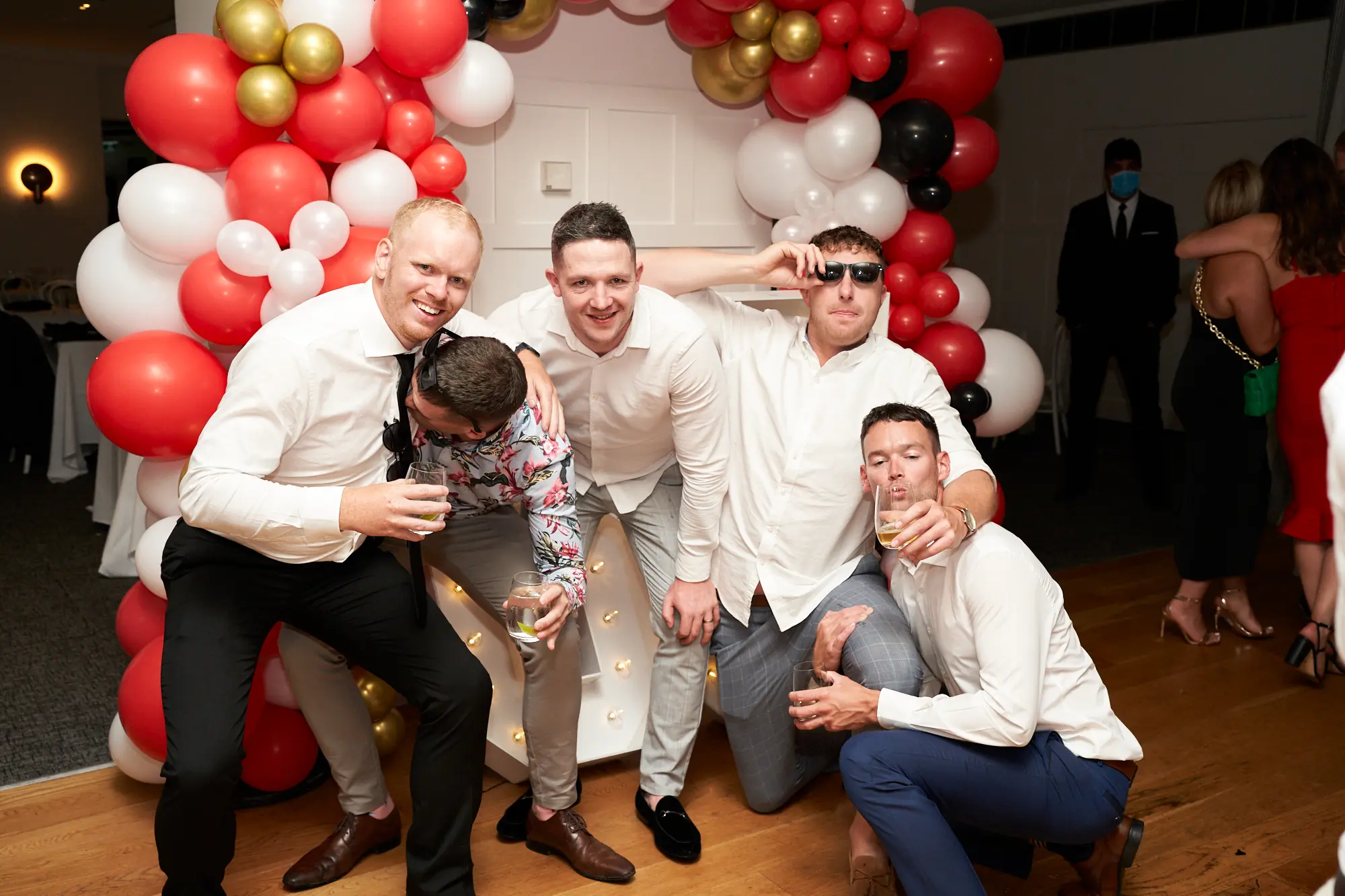 garryowens 65th anniversary ball five friends group Five men pose together indoors during Garryowen’s 65th Anniversary Ball at the Royal Melbourne Yacht Squadron, smiling broadly and holding drinks. Four stand in the back row while one crouches in front, all dressed in formal or cocktail attire. Behind them, a large festive balloon arch in red, white, gold, and black frames the group, with white-paneled walls and mingling guests visible in the distance. The polished hardwood floor reflects the warm ambient lighting. The scene captures camaraderie, celebration, and shared joy in a lively, sophisticated, and memorable gala atmosphere.