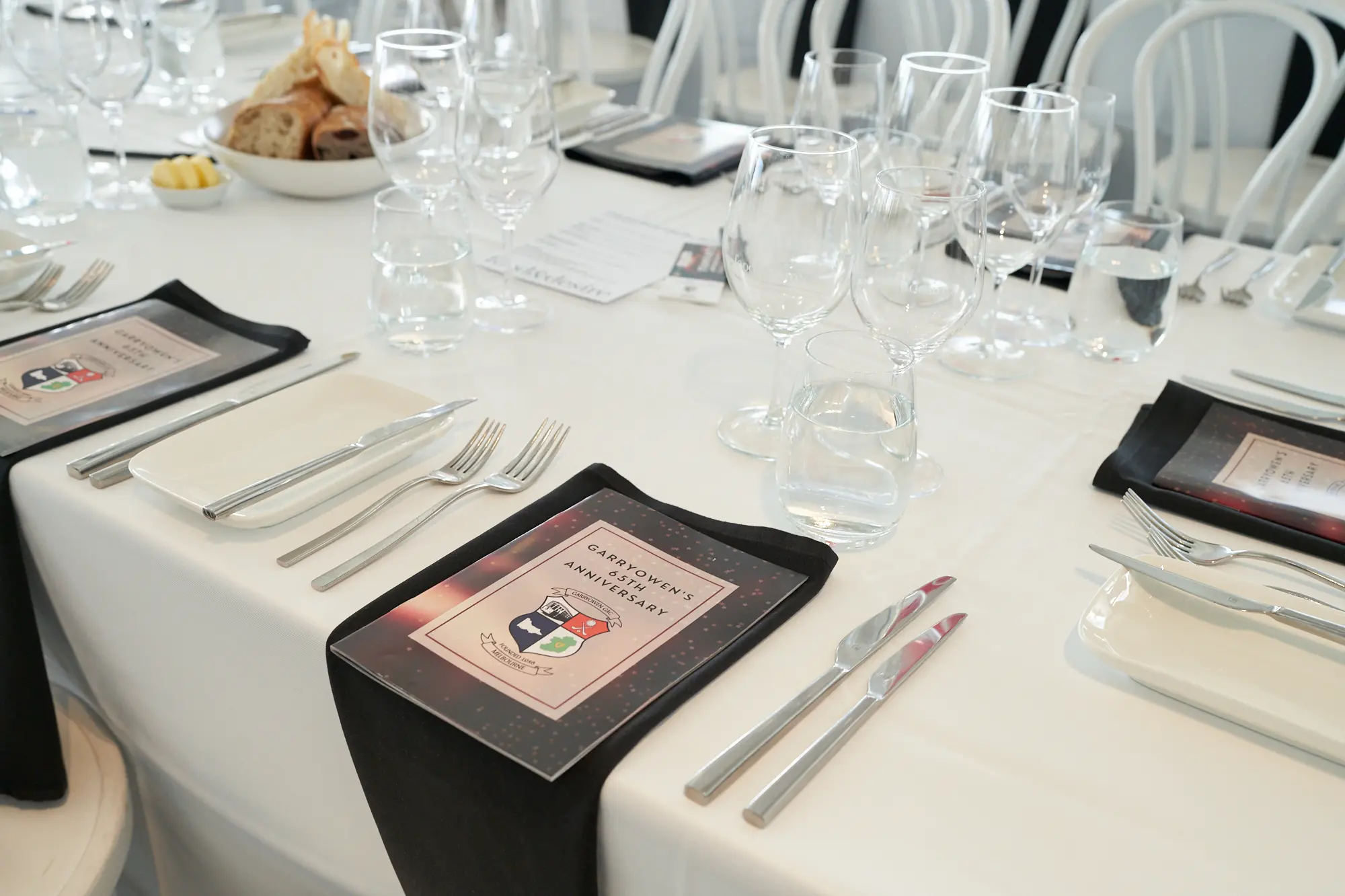 garryowens 65th anniversary ball formal table setting closeup A close-up of an elegantly set dinner table prepared for Garryowen’s 65th Anniversary Ball shows polished cutlery, multiple wine and water glasses, neatly folded black napkins, and personalised anniversary programs with gold and red accents. The bright event space in the background features rows of white chairs and tables covered in crisp white linens, softly blurred to keep focus on the detailed place setting. The scene captures a refined sense of anticipation, showcasing the meticulous preparation, chic black-and-white theme, and sophisticated atmosphere awaiting guests at this formal awards night celebration.