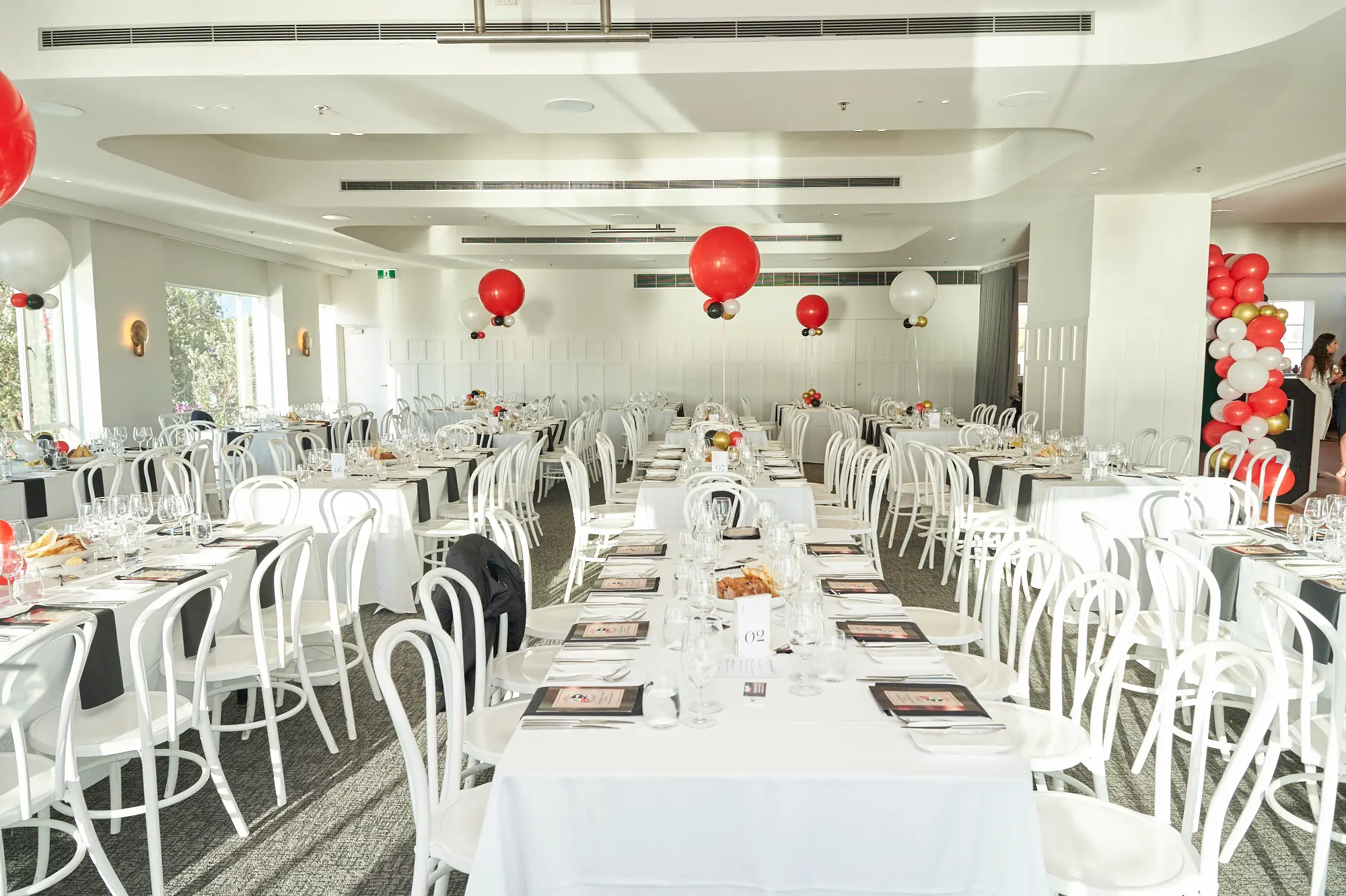 garryowens 65th anniversary ball formal venue overview decor setup A spacious ballroom is fully prepared for Garryowen’s 65th Anniversary Ball, featuring long dining tables dressed in white linens, black runners, polished glassware, and neatly arranged white chairs. Bright, even lighting highlights the crisp black-and-white theme, while vibrant red, white, gold, and black balloons— including a large balloon arch—add celebratory flair. The modern event space includes curved ceiling details, white walls, and windows that softly illuminate the room. The meticulously arranged décor and clean, elegant presentation create an atmosphere of sophistication and anticipation before guests arrive for the awards night celebration.