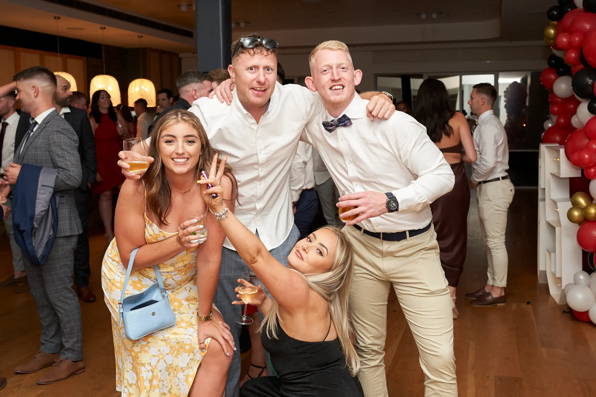 garryowens 65th anniversary ball four friends indoor Four friends pose energetically together at Garryowen’s 65th Anniversary Ball at the Royal Melbourne Yacht Squadron. Two men stand behind two women, all smiling at the camera. The crouching woman in the foreground holds a champagne glass and flashes a peace sign, while the men link arms in a friendly gesture. The indoor venue features polished hardwood floors, white walls, modern track lighting, and a festive balloon arch in red, black, gold, and white. Other guests mingle in the background, adding to the lively atmosphere. The image captures celebration, camaraderie, and joyful social interaction in a sophisticated setting.