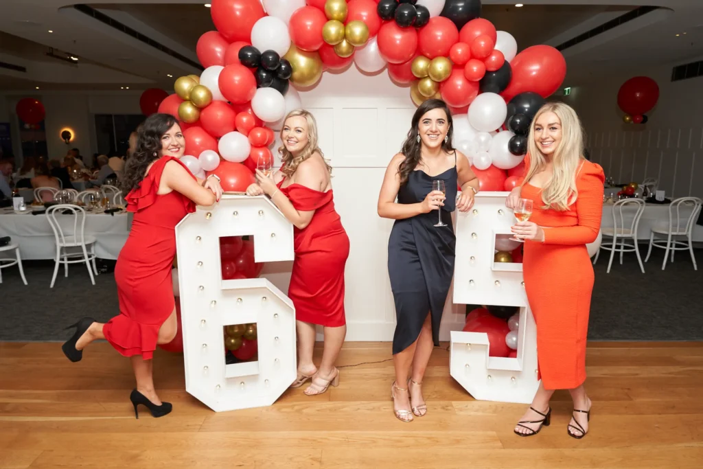 svg%3E Four elegantly dressed women pose around large illuminated “6” and “5” numbers during Garryowen’s 65th Anniversary Ball at the Royal Melbourne Yacht Squadron. Smiling brightly and holding glasses for a toast, they exude glamour and celebration. Behind them, a festive balloon arch in red, gold, black, and white frames the milestone “65,” while polished wooden floors and elegantly set tables highlight the formal indoor venue. This image captures the chic, joyous, and sociable atmosphere of the awards night, showcasing friends commemorating the special occasion with style, warmth, and lively engagement.