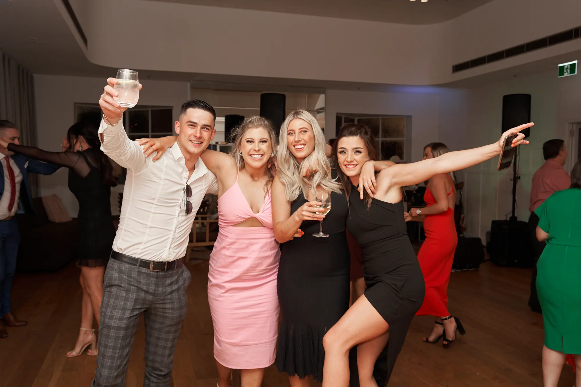 garryowens 65th anniversary ball friends celebration yacht squadron Four friends pose together at Garryowen’s 65th Anniversary Ball at the Royal Melbourne Yacht Squadron. Two men and two women stand closely, smiling brightly at the camera. One man raises a glass in a toast, while the woman on the right extends an arm backward and holds the waist of her friend. Other guests mingle in the softly lit, elegant indoor venue with polished hardwood floors. Warm ambient lighting highlights the joyful expressions, dynamic poses, and lively social energy, capturing a celebratory moment filled with camaraderie, excitement, and the sophistication of a formal gala event.