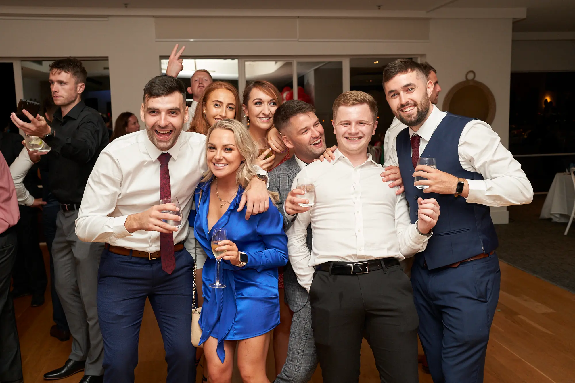 garryowens 65th anniversary ball group celebration Nine friends, seven men and two women, pose together indoors during Garryowen’s 65th Anniversary Ball, holding drinks and smiling brightly. Some raise hands in celebration, while one man flashes a peace sign. The group stands closely, arms around shoulders, dressed in formal and cocktail attire. The polished hardwood floor reflects ambient lighting, with large glass windows and a distant bar area visible. Clean white walls and a dark column frame the space. The scene captures lively camaraderie, shared joy, and an energetic, celebratory atmosphere at a sophisticated evening gala event.