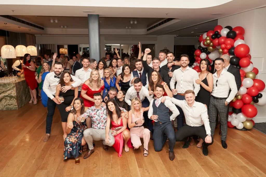 garryowens 65th anniversary ball group photo royal melbourne yacht squadron st kilda A large group of guests pose together during Garryowen’s 65th Anniversary Ball at the Royal Melbourne Yacht Squadron in St Kilda. Around thirty well-dressed attendees gather in multi-level rows, smiling, laughing, and holding drinks as they celebrate the milestone event. The polished hardwood floors, warm lighting, and vibrant red, black, gold, and white balloon arch create a festive atmosphere, while more guests mingle near the bar in the background. This lively group portrait captures the joyful energy, camaraderie, and elegance of the anniversary awards night.