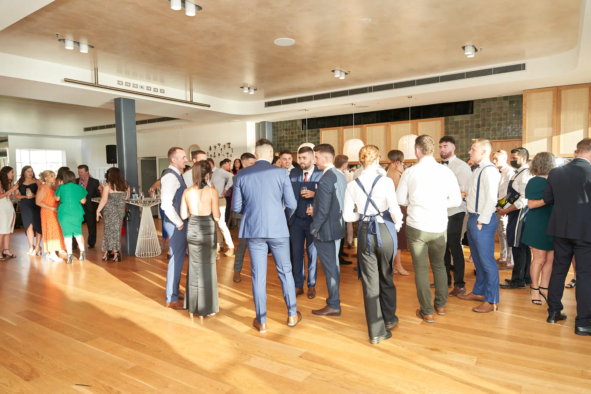 garryowens 65th anniversary ball guest arrivals socialising reception Guests arrive for Garryowen’s 65th Anniversary Ball, gathering in lively groups throughout a bright waterfront venue. Men in suits form the central cluster, chatting enthusiastically while others mingle nearby with drinks in hand. Floor-to-ceiling windows fill the room with natural light and offer views of the water, contrasting with the warm interior tones of hardwood floors and modern decor. The elegant attire, animated conversations, and welcoming atmosphere capture the excitement and energy of the evening’s opening moments, reflecting the anticipation of a formal anniversary celebration and awards night.