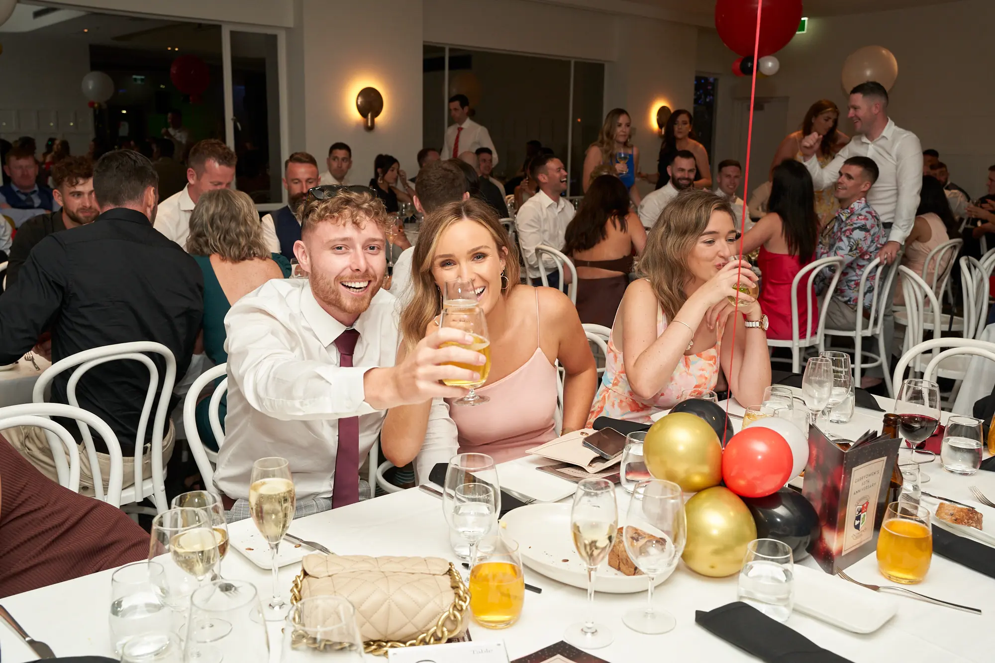 garryowens 65th anniversary ball guest toast celebration Guests celebrate joyfully at Garryowen’s 65th Anniversary Ball, seated at a formal dining table. A man and woman clink glasses in a cheerful toast, smiling widely at each other, while another woman nearby laughs, hands near her face. The table is adorned with drinks, glassware, and decorative balloons, set against a polished indoor venue with white walls, warm lighting, and a festive red, black, gold, and white balloon arch. Other guests mingle and enjoy the festivities in the background, creating a lively, sophisticated, and warm gala atmosphere filled with shared happiness and excitement.