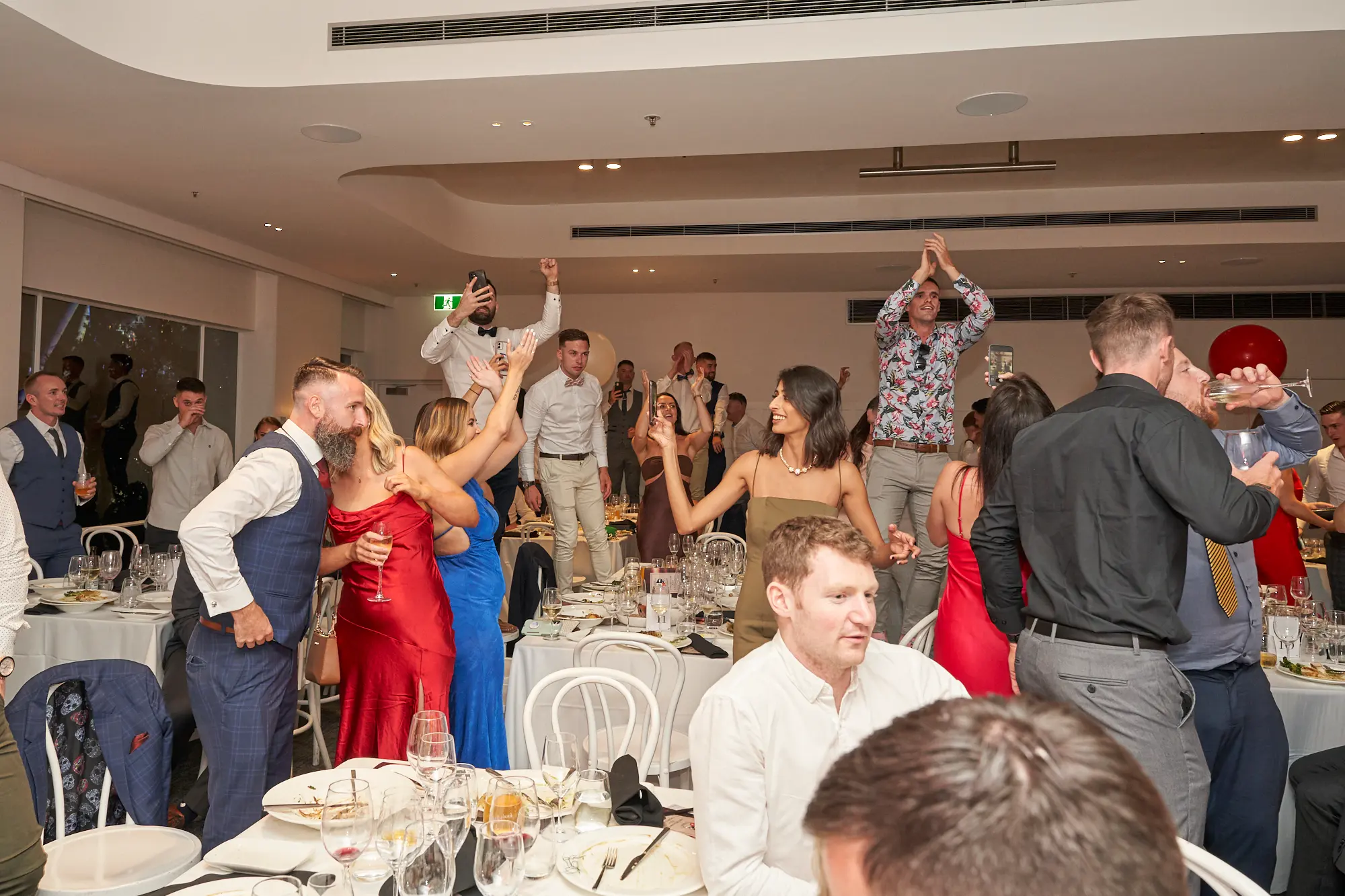 garryowens 65th anniversary ball guests cheering tables A lively group of guests celebrate at Garryowen’s 65th Anniversary Ball at the Royal Melbourne Yacht Squadron. Dressed in formal and cocktail attire, two men stand on a dining table with arms raised, while a woman in red embraces another in blue. Other guests hold drinks and cheer amid white tablecloth-covered tables and white chairs. Red balloons float above the polished hardwood floor of the modern, bright event space. The dynamic poses, energetic gestures, and joyful expressions capture the electric, high-energy atmosphere of a festive awards night and milestone anniversary gala.