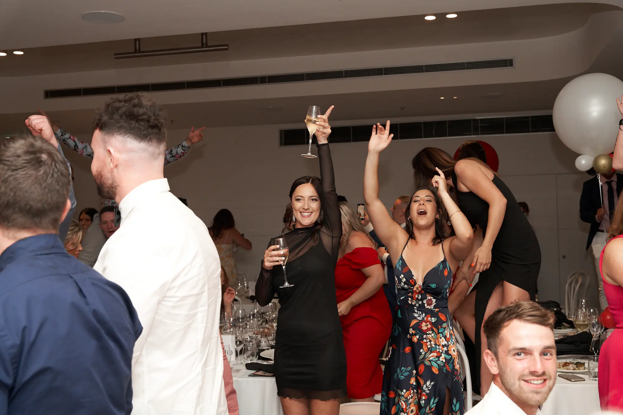 garryowens 65th anniversary ball guests dancing cheering Guests celebrate energetically at Garryowen’s 65th Anniversary Ball at the Royal Melbourne Yacht Squadron. Two women in the mid-ground cheer with arms raised—one in a black mesh dress holding champagne, the other in a floral dress shouting joyfully—while a man on the left dances with arms up. Other guests smile, laugh, and hold drinks across the polished hardwood floor, surrounded by white tablecloth-draped tables and white chairs. A large white balloon decorates the space, and a bar is visible in the distance. The dynamic poses and festive energy capture the electric, high-spirited atmosphere of a lively awards night and gala celebration.