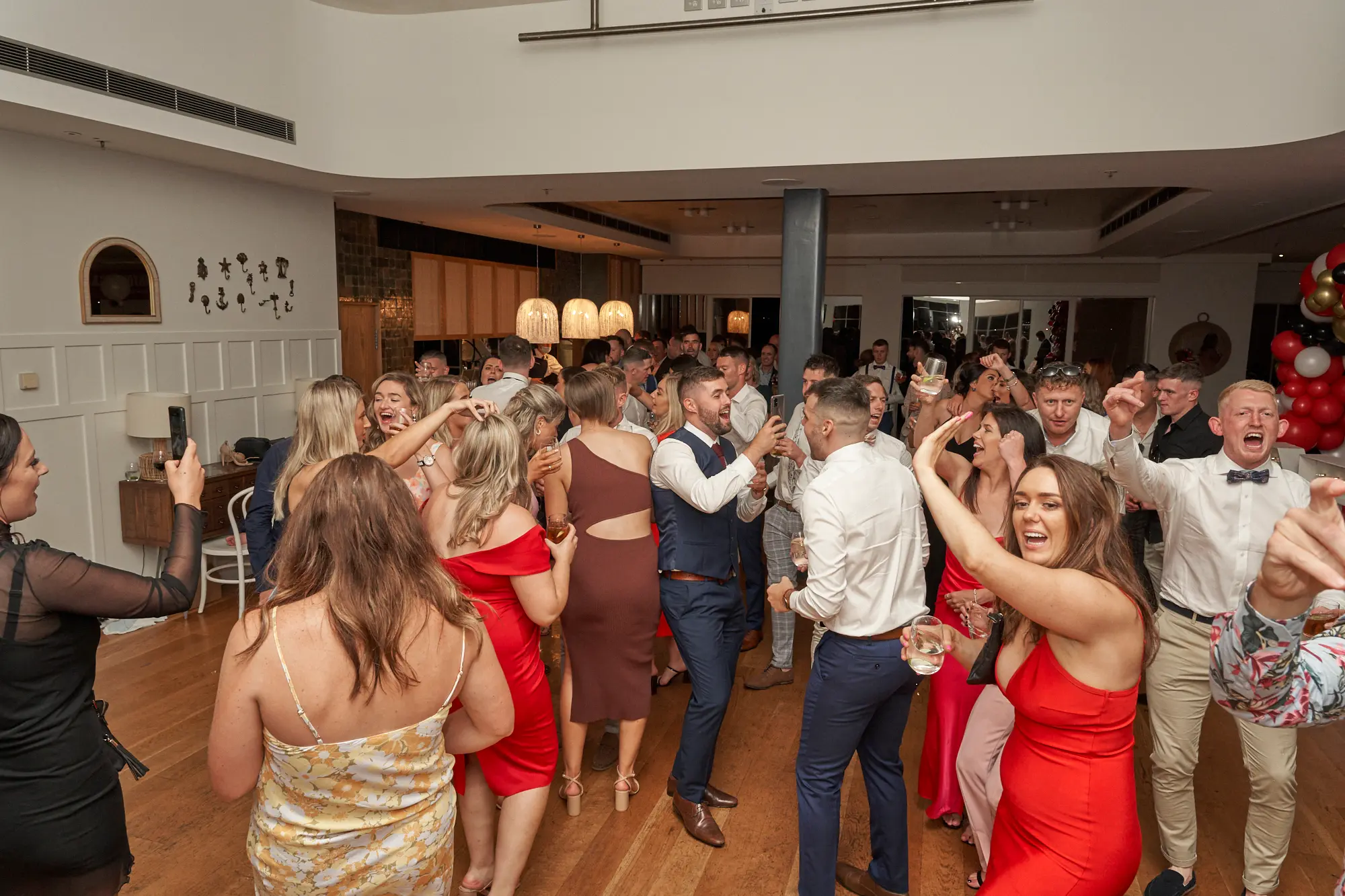garryowens 65th anniversary ball guests dancing A lively scene at Garryowen’s 65th Anniversary Ball at the Royal Melbourne Yacht Squadron captures guests dancing, cheering, and posing for photos. Two women in the mid-ground raise their arms in excitement, one holding a glass, while a man on the right cheers enthusiastically. Guests in formal and cocktail attire hold drinks and engage with each other amid polished hardwood floors, white walls, a large festive balloon arch in red, black, gold, and white, and softly lit dining tables, creating a vibrant, high-energy atmosphere full of joy, celebration, and shared happiness.