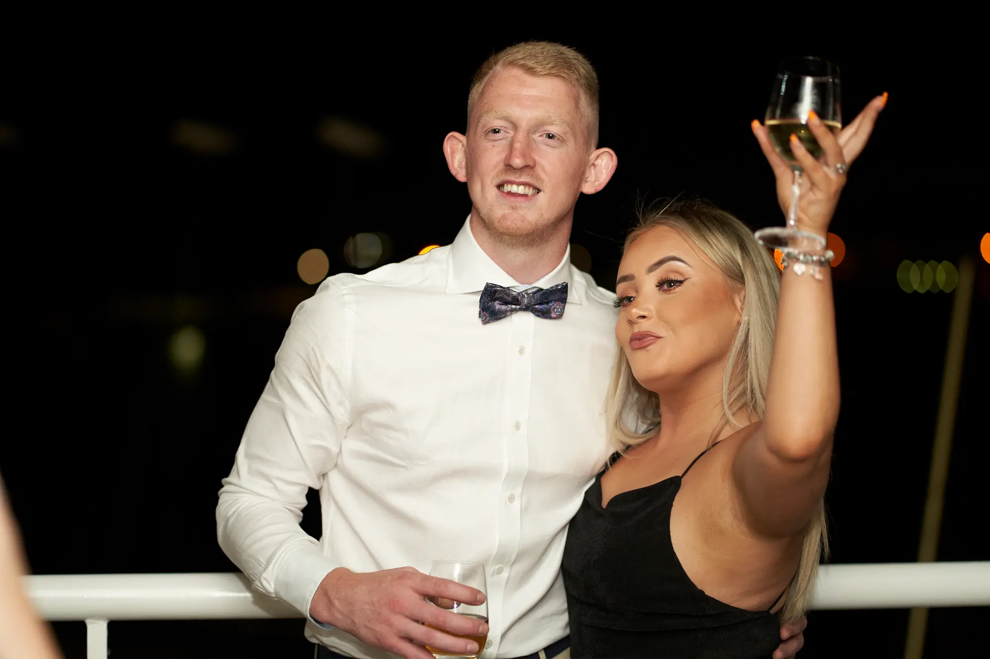 garryowens 65th anniversary ball harbour room night balcony couple A couple poses joyfully on the Harbour Room balcony at night during Garryowen’s 65th Anniversary Ball. The man in a white shirt and patterned bow tie smiles slightly off-camera, holding a glass, while the woman in a black slip dress raises a champagne glass mid-cheer. The white balcony railing frames them against a dark sky and distant city lights across the bay. Their animated expressions and celebratory pose convey excitement, shared happiness, and nighttime elegance, capturing the lively, sophisticated atmosphere of the gala’s evening festivities.
