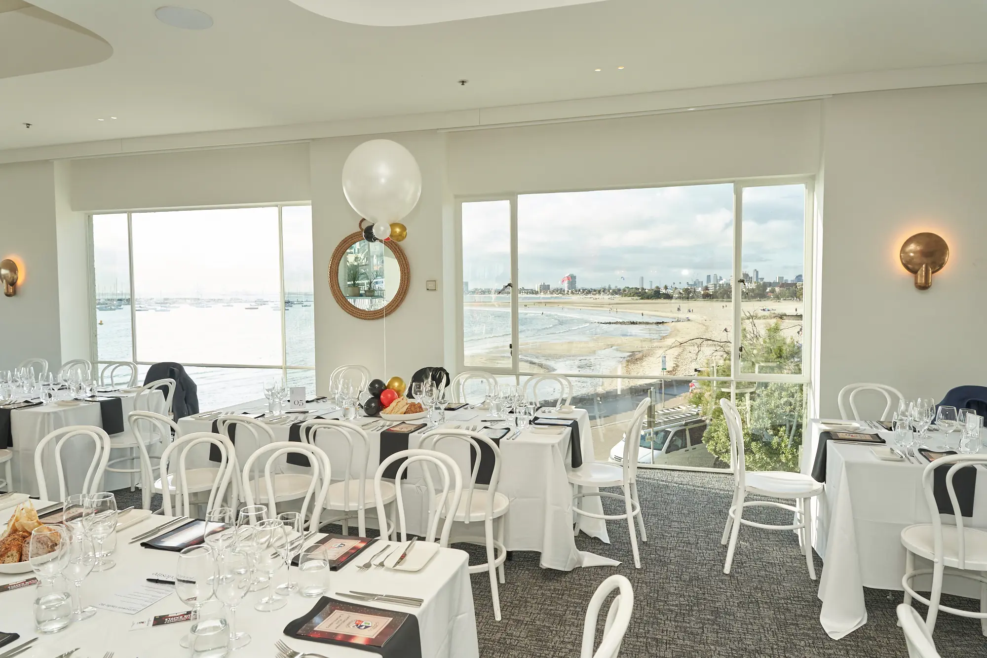 garryowens 65th anniversary ball harbour room venue decor setup The Harbour Room at the Royal Melbourne Yacht Squadron is elegantly prepared for Garryowen’s 65th Anniversary Ball, with dining tables dressed in white linens and black-and-white table settings accented with touches of red and gold. Floor-to-ceiling windows reveal panoramic views of St Kilda Beach and Port Phillip Bay, with sand, water, and distant boats under a soft, cloudy sky. The bright, modern interior, polished light oak floors, and decorative balloon clusters convey sophisticated anticipation, capturing the refined elegance and serene beauty of the venue before guests arrive for the milestone awards night celebration.