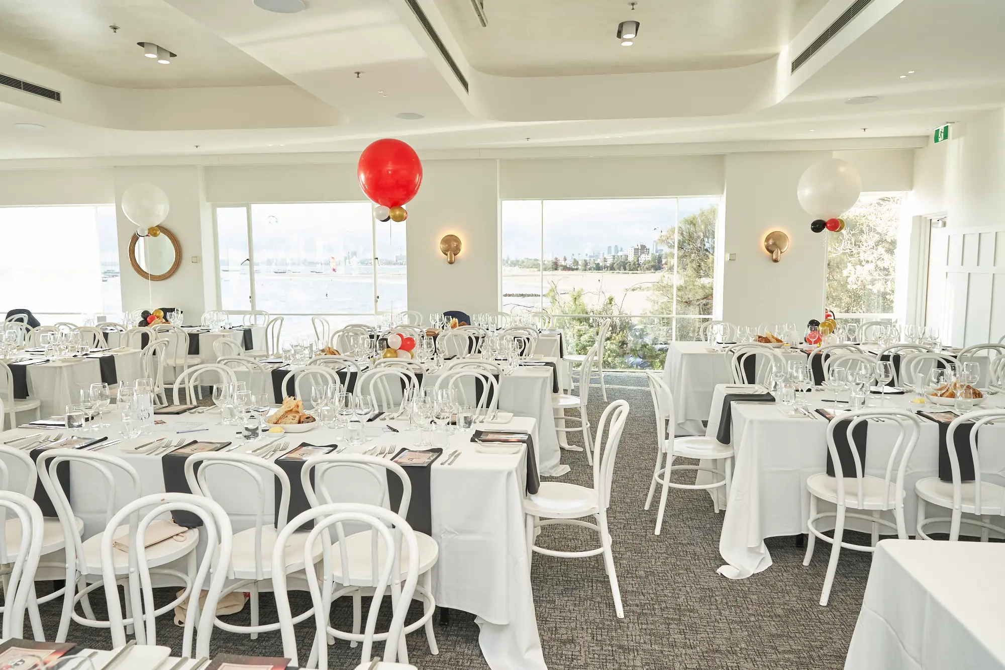 garryowens 65th anniversary ball harbour room wide venue overview A wide view of the Harbour Room at the Royal Melbourne Yacht Squadron shows the venue fully prepared for Garryowen’s 65th Anniversary Ball. Floor-to-ceiling windows flood the space with natural light and offer sweeping views of the bay, trees, and coastal surroundings. Inside, rows of tables dressed in crisp white tablecloths with black runners are paired with neatly arranged white bentwood chairs, each table set with polished glassware and cutlery. Festive red and white balloons add vibrant pops of color. The bright, modern room conveys refined elegance and calm anticipation before guests arrive for the celebration.