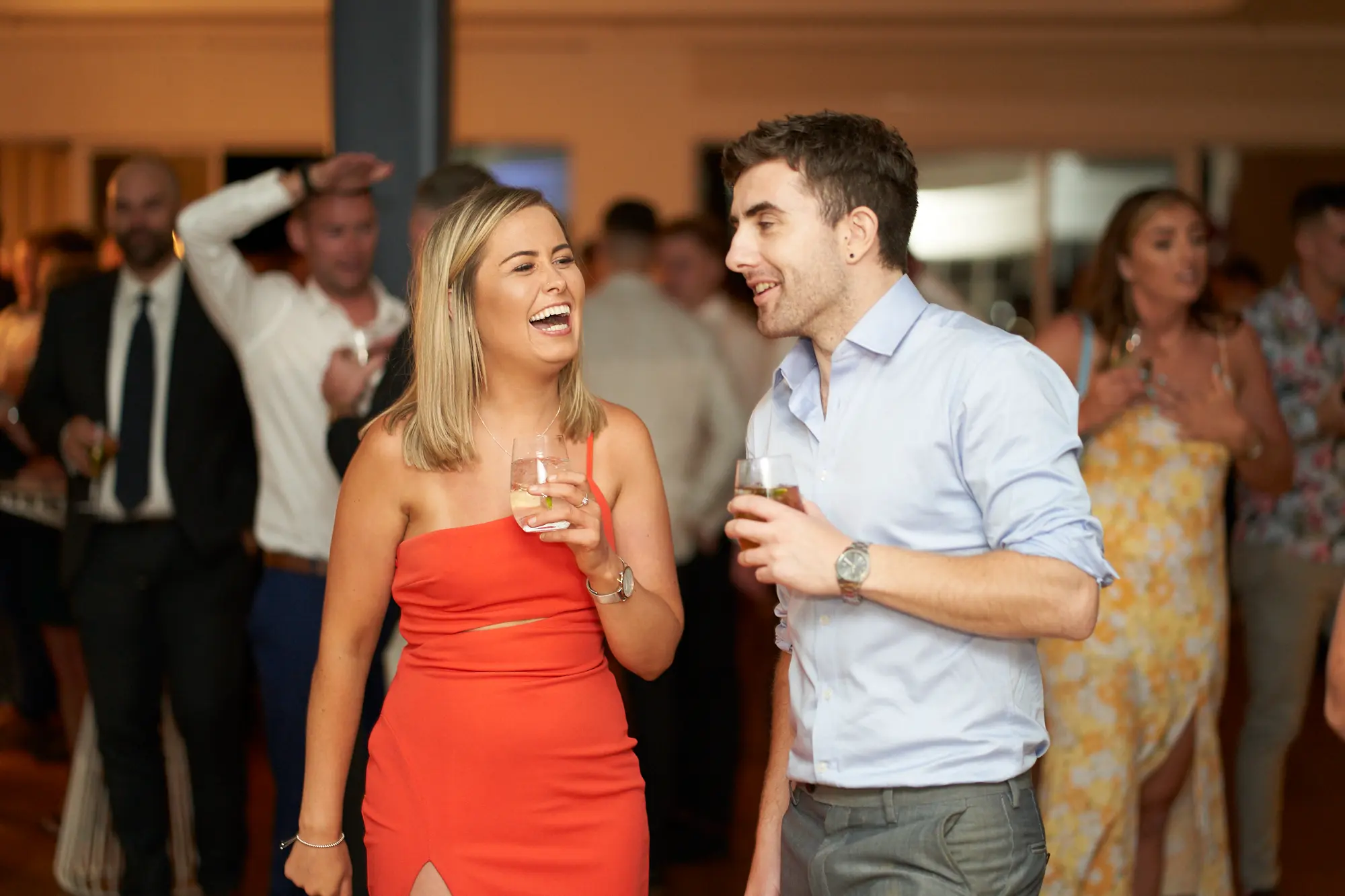 garryowens 65th anniversary ball indoor candid laughter Two guests share a joyful moment at Garryowen’s 65th Anniversary Ball, with the woman in a bright orange one-shoulder dress laughing heartily while holding a drink, and the man in a light blue shirt smiling back, also holding a drink. They stand in a polished indoor venue filled with formally dressed guests, softly lit by warm overhead lights. The shallow depth of field emphasizes their animated interaction while showing a lively background of mingling attendees, capturing a cheerful, social, and celebratory atmosphere during the gala event.