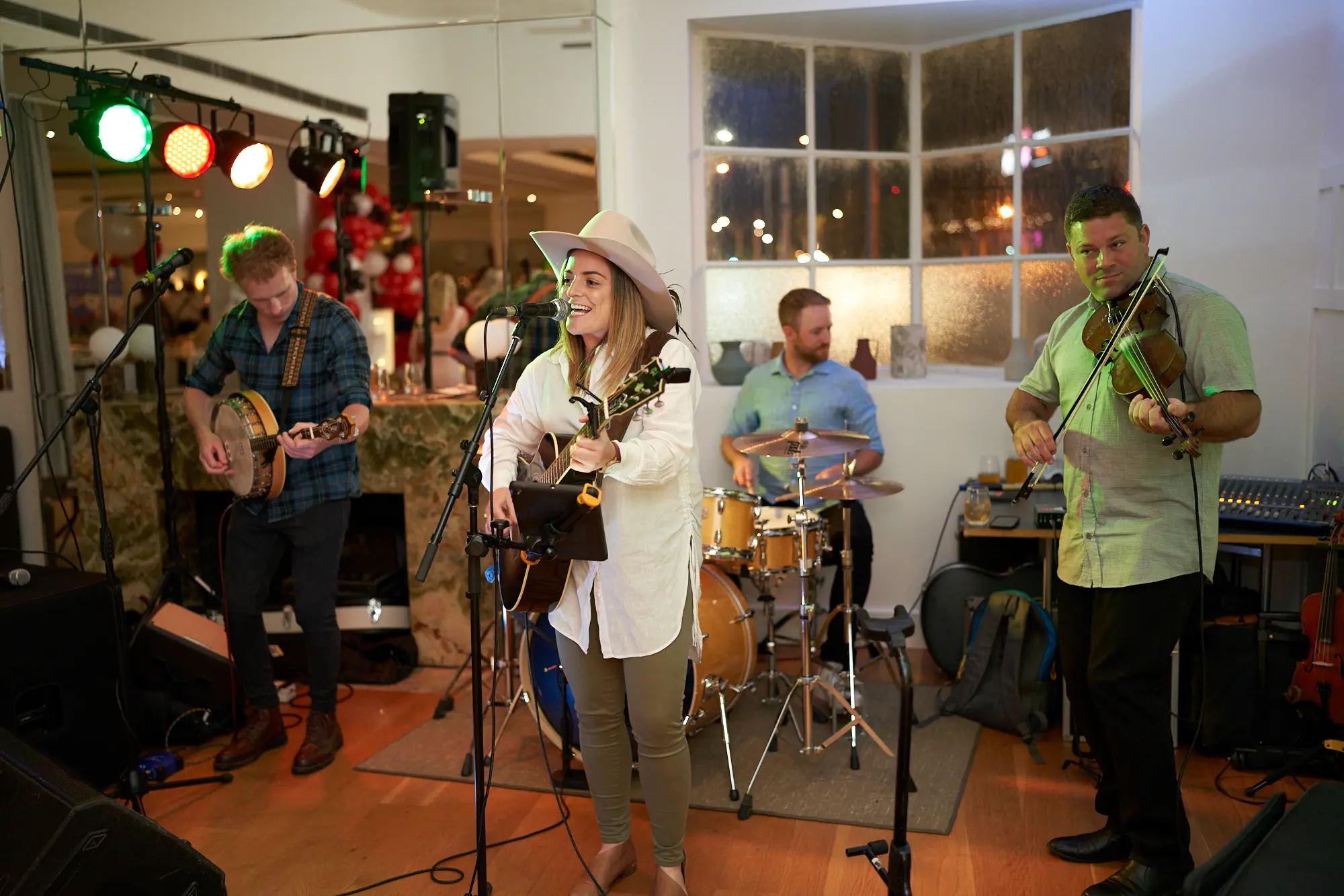 garryowens 65th anniversary ball live band performance A four-piece band performing live at Garryowen’s 65th Anniversary Ball at the Royal Melbourne Yacht Squadron. A male banjo player strums intently in the foreground, while a female guitarist and singer wearing a hat plays and sings into a microphone. Behind them, a drummer and a standing violinist add to the performance. Stage lighting in red and green illuminates the polished hardwood floor and white-walled venue. Professional audio equipment surrounds the band, while guests mingle in the softly lit background. The scene captures the vibrant energy and lively atmosphere of live entertainment during the formal awards night.