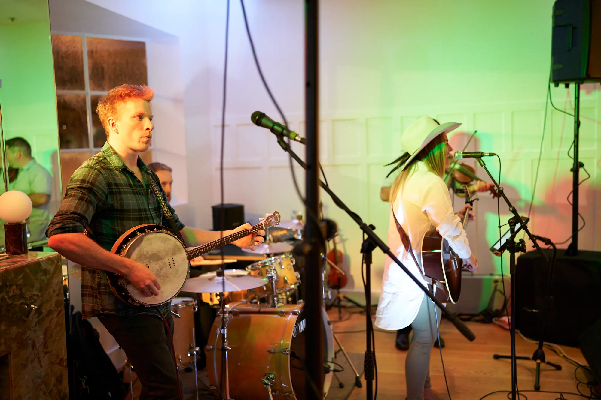 garryowens 65th anniversary ball live music performance Musicians performing live at Garryowen’s 65th Anniversary Ball at an indoor event venue. A male banjo player strums in profile while a female guitarist and singer wearing a white hat plays and sings into a microphone. Behind them, a drummer and violinist add to the ensemble. Stage lighting in vibrant green highlights the polished hardwood floor and elegant walls. Guests mingle near a bar in the background. The scene captures the energy, engagement, and lively atmosphere of the awards night, showcasing live entertainment as a central part of the celebration.