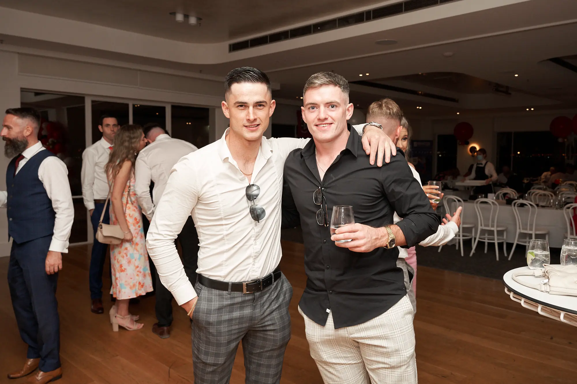 garryowens 65th anniversary ball royal melbourne yacht squadron guest embrace Two men share a joyful embrace at Garryowen’s 65th Anniversary Ball at the Royal Melbourne Yacht Squadron, smiling directly at the camera. The man in a black shirt and white trousers holds a glass of amber-colored drink while wrapping his arm around his friend in a white shirt and grey plaid trousers, who has his hand in his pocket. Sunglasses are hooked onto both shirts. Behind them, guests mingle near white-draped tables, festive red balloons float nearby, and warm ambient lighting illuminates the elegant, lively indoor venue, capturing the excitement and camaraderie of the celebration.