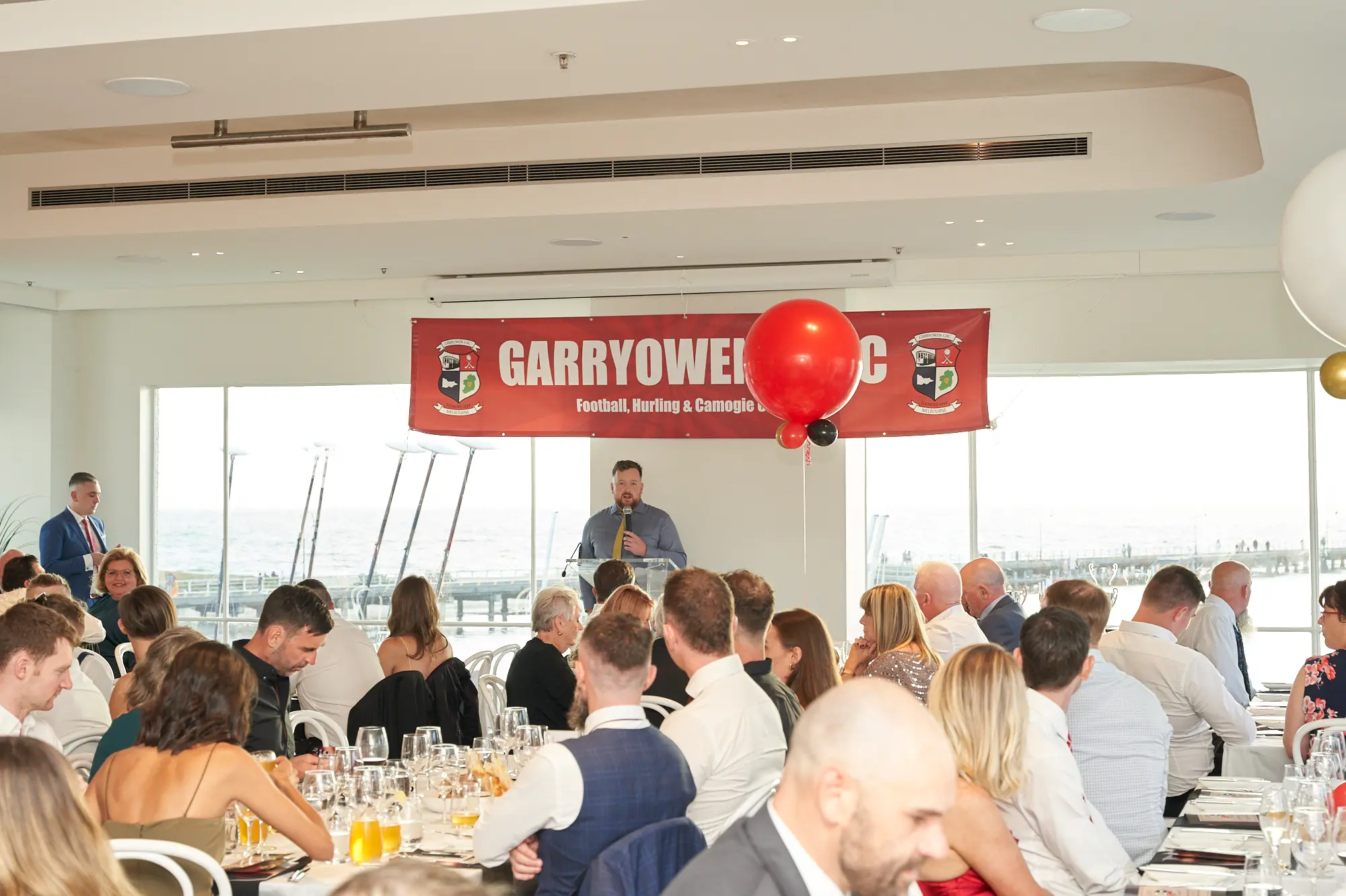 garryowens 65th anniversary ball speaker awards ceremony A speaker addresses guests from a central podium during Garryowen’s 65th Anniversary Ball and awards night. Seated attendees at round tables listen attentively, while some converse quietly with table companions. Behind the speaker, a large red banner displays “GARRYOWEN” and crests for Football, Hurling & Camogie, with a festive red balloon nearby. Floor-to-ceiling windows reveal waterfront views outside, and the bright, modern venue features polished floors and well-set tables. The image captures a formal yet celebratory atmosphere, combining the focus of the official speeches and awards with the lively, engaged energy of guests enjoying the milestone evening.