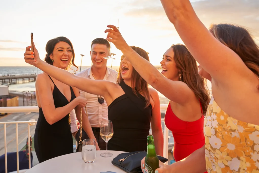 svg%3E A group of five friends in elegant evening attire share a joyful, candid moment on a balcony during Garryowen’s 65th Anniversary Ball at the Royal Melbourne Yacht Squadron. Four women and one man gather around a smartphone, taking a selfie while laughing and raising their arms in celebration. Behind them, the golden-hour sky casts warm light over the sparkling bay and a distant pier. Their raised glasses, bright smiles, and lively gestures capture the intimate, festive, and celebratory atmosphere of this milestone event, showcasing friendship, connection, and the unforgettable energy of the evening.