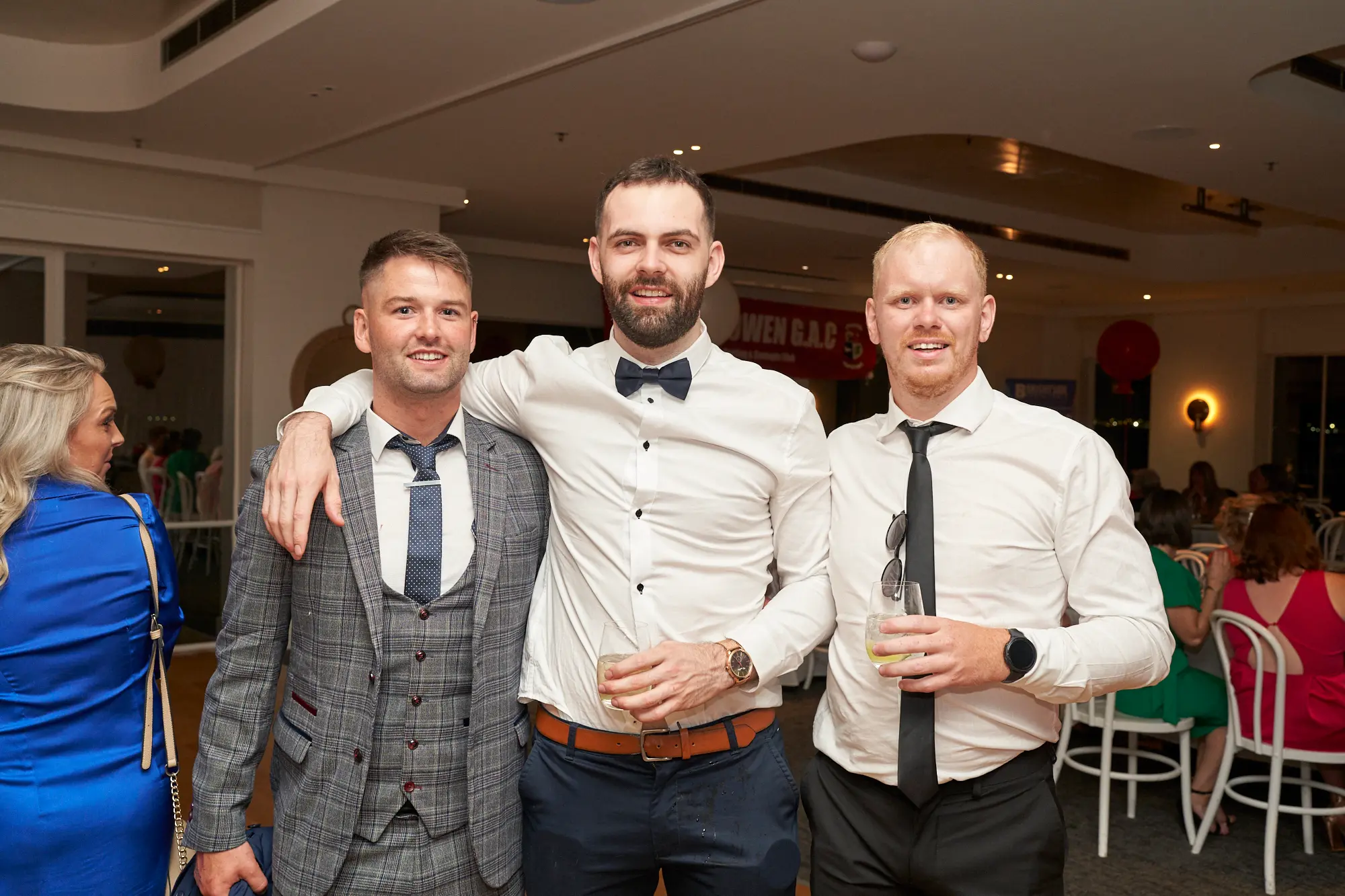 garryowens 65th anniversary ball three friends cocktail Three men pose closely together indoors at Garryowen’s 65th Anniversary Ball, smiling warmly and holding drinks. The man in the center has his arms around the shoulders of the men beside him, all dressed in formal or cocktail attire. Behind them, red balloons and a partial banner reading “WEN G.A.C” add a festive touch, while other guests mingle at tables and near the bar. The polished hardwood floor and light-coloured walls reflect warm ambient lighting. The scene captures friendship, celebration, and lively social interaction in a sophisticated and joyful gala environment.