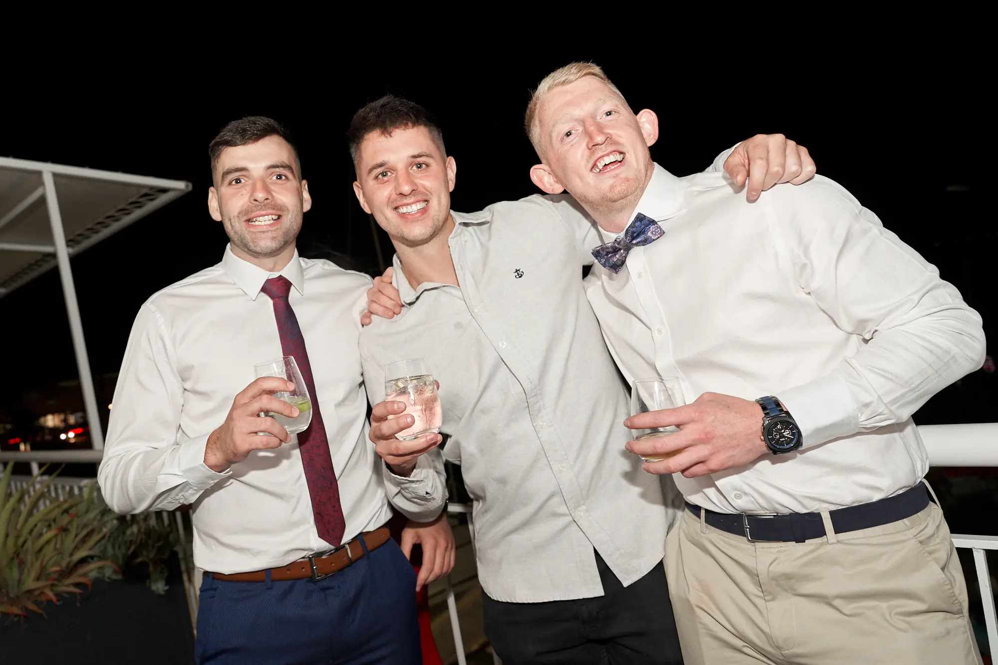 garryowens 65th anniversary ball three men celebration Three men pose together on an outdoor deck at night during Garryowen’s 65th Anniversary Ball, smiling broadly and holding glasses with lime garnishes. The man on the right has his arm around the center man, while all wear formal attire, including a burgundy tie, grey open-collar shirt, and white shirt with patterned bow tie. White railings frame the deck, and subtle ambient lighting highlights their joyful expressions against the dark nighttime backdrop. A small planter with green foliage is visible to the left. The scene captures camaraderie, celebration, and an elegant, lively atmosphere at a sophisticated evening gala event.