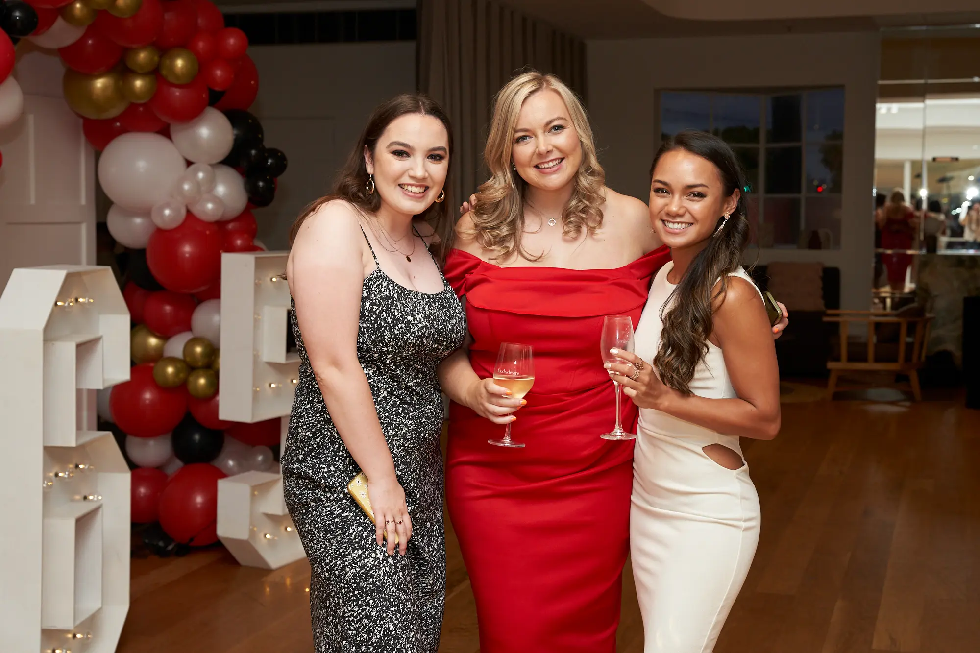 garryowens 65th anniversary ball three women group portrait Three elegantly dressed women pose together at Garryowen’s 65th Anniversary Ball, smiling warmly at the camera. The woman on the left holds a glass of amber drink, the center wears a red off-shoulder dress, and the right wears a white cut-out dress, both holding wine glasses. Behind them, a festive balloon arch in red, black, gold, and white and illuminated marquee numbers set a celebratory backdrop, while guests mingle near a softly lit bar. The polished hardwood floor, subtle architectural details, and warm ambient lighting create a sophisticated, lively, and joyful gala atmosphere.