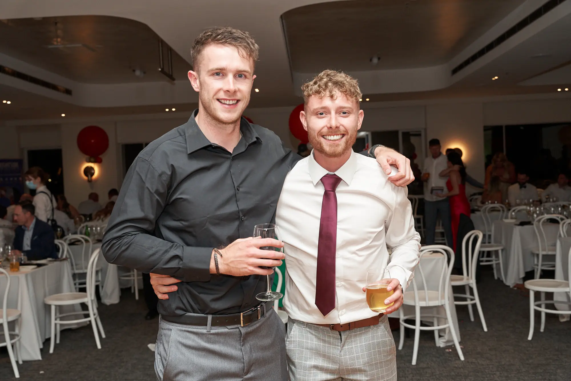garryowens 65th anniversary ball two men celebration Two men share a warm embrace at Garryowen’s 65th Anniversary Ball at the Royal Melbourne Yacht Squadron, smiling broadly at the camera. The man on the right wears a white shirt, burgundy tie, and plaid trousers while holding a beer, and the man on the left wears a dark grey shirt and light grey trousers, holding a wine glass. Surrounded by formally set tables, mingling guests, and festive red balloons, the elegant indoor venue exudes a lively, celebratory atmosphere. Their relaxed, friendly body language conveys camaraderie, excitement, and shared enjoyment during a sophisticated social gathering.