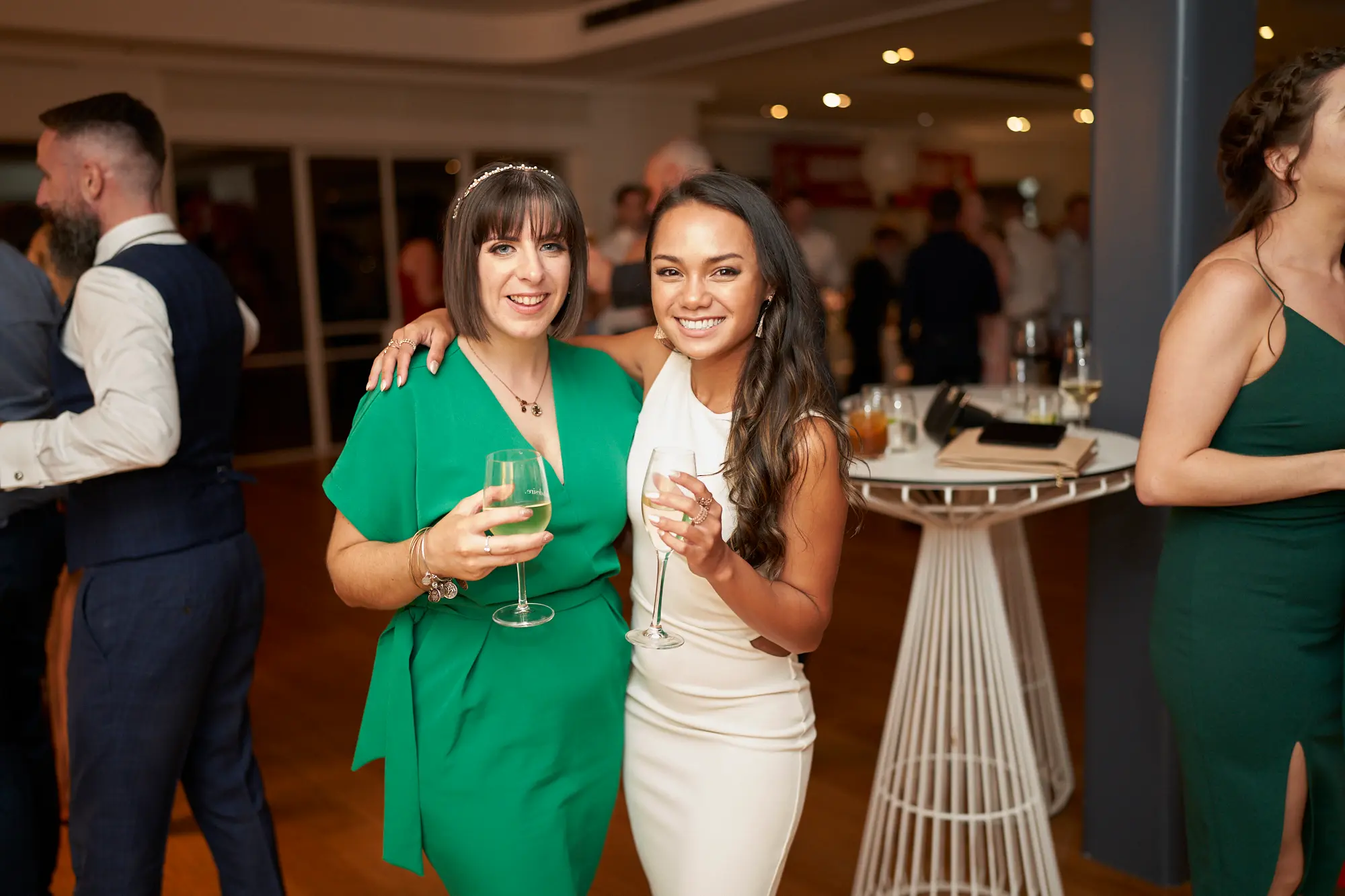 garryowens 65th anniversary ball two women celebration Two women pose side-by-side at Garryowen’s 65th Anniversary Ball at the Royal Melbourne Yacht Squadron, smiling warmly at the camera. The woman on the left wears an elegant green dress, holding a light-colored drink, while the woman on the right wears a white dress, holding a glass of wine. Behind them, a man in a waistcoat and mingling guests add depth to the sophisticated indoor venue. The polished hardwood floor, modern high-top tables, warm ambient lighting, and formally dressed attendees create a lively, celebratory atmosphere, capturing friendship, joy, and a festive social moment at a milestone gala event.