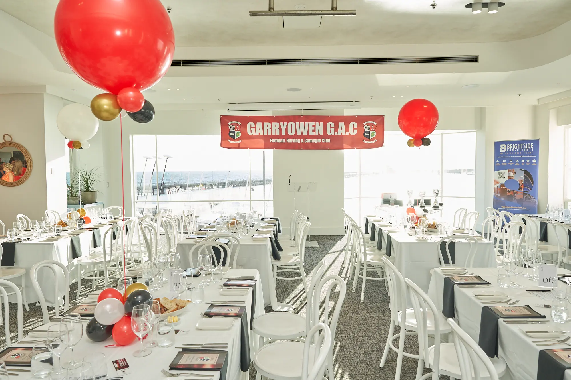 garryowens 65th anniversary ball venue decor harbour room banner The Harbour Room at the Royal Melbourne Yacht Squadron is elegantly prepared for Garryowen’s 65th Anniversary Ball and awards night. Dining tables are dressed in white linens with black-and-white table settings, while red, white, gold, and black balloon arrangements add festive color. A large red banner reading “GARRYOWEN G.A.C” hangs above floor-to-ceiling windows that showcase a panoramic view of the outdoor deck and bay. The bright, modern interior, light-colored floors, and natural sunlight create a sophisticated, inviting atmosphere, capturing the venue’s stylish decor, serene elegance, and anticipation before guests arrive for the milestone celebration.